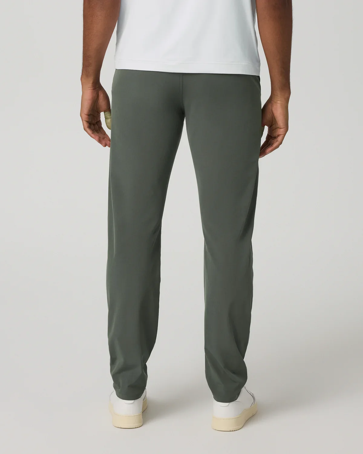 A person wearing dark green pants, a white shirt, and white shoes stands facing away from the camera against a plain light background, ready to pick out the best golf travel bags for their next adventure.