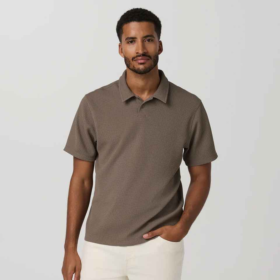 A man with short hair and a beard wears a textured brown Vuori polo shirt and light-colored pants, standing against a plain light gray background. He has one hand in his pocket and a neutral expression—perfect Father's Day golf apparel.