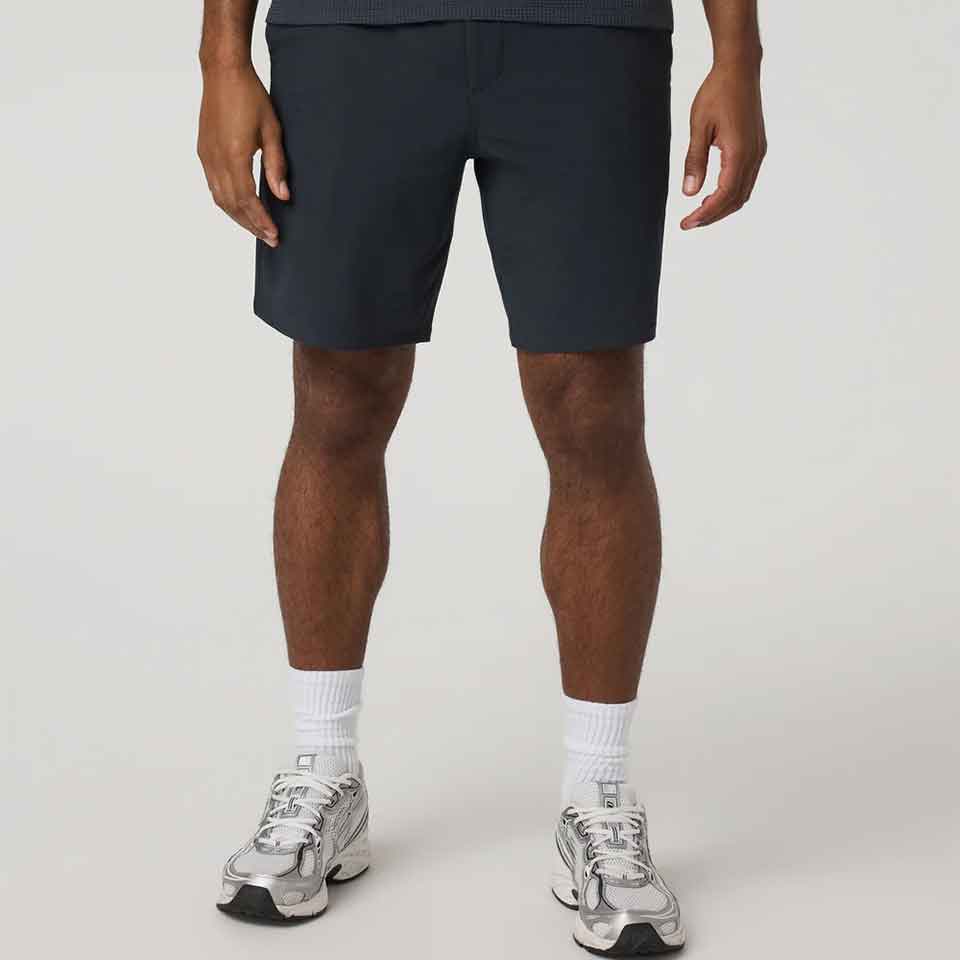 A person stands against a plain background, wearing Vuori dark shorts, white crew socks, and silver athletic sneakers—their upper body is not visible in the image.