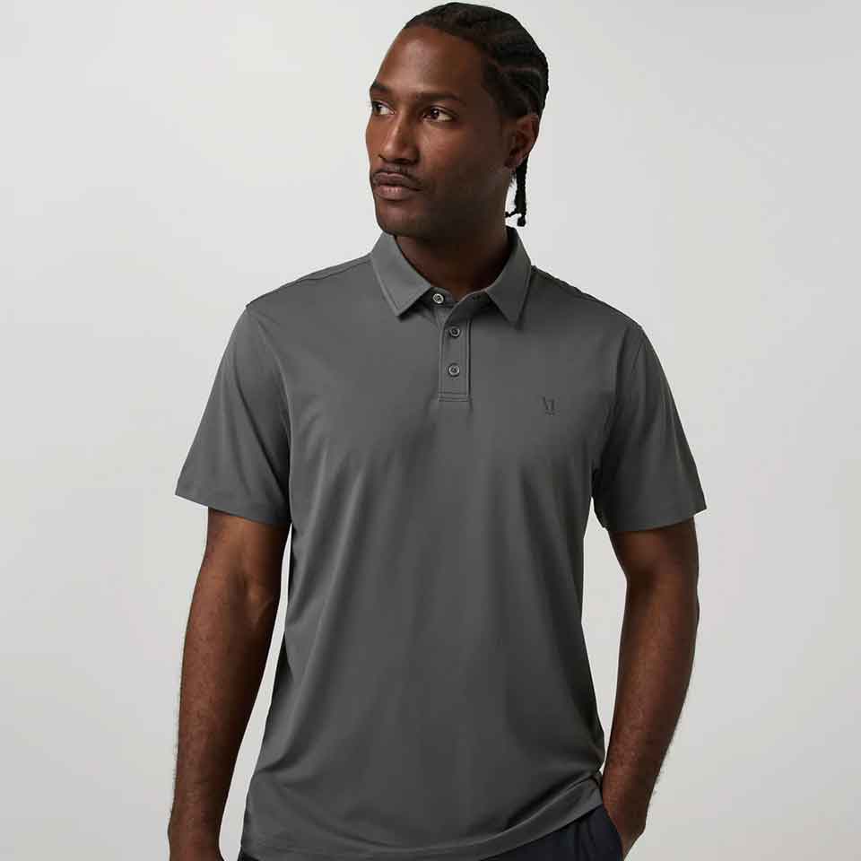 A man with braided hair wears a short-sleeve, gray Vuori polo shirt—perfect golf apparel for Father’s Day—as he stands against a plain, light background, looking slightly to his left with one hand in his pocket.