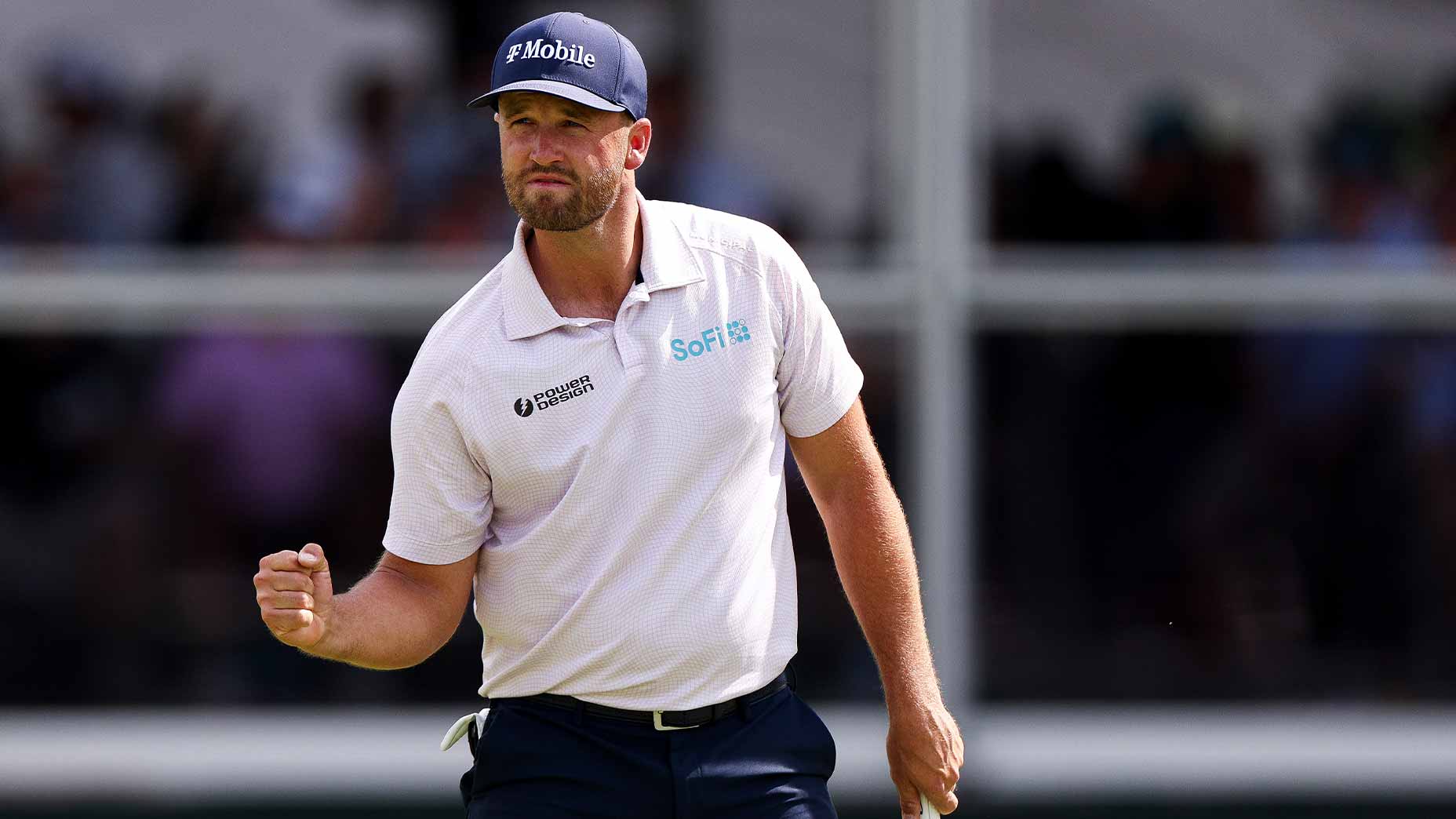 A golfer, inspired by mental-game coach techniques to re-frame thoughts, wearing a navy cap and light polo shirt pumps his fist in celebration on the course, with blurred spectators in the background.