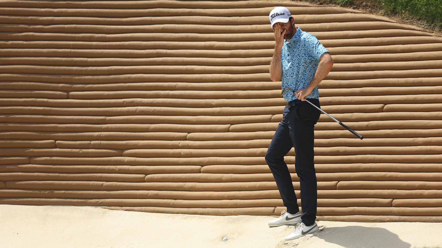 A golfer stands in a sand bunker, holding a golf club in one hand and covering his face with the other, appearing frustrated. He wears a patterned shirt, dark pants, white shoes, and a cap, with a textured bunker wall behind him.
