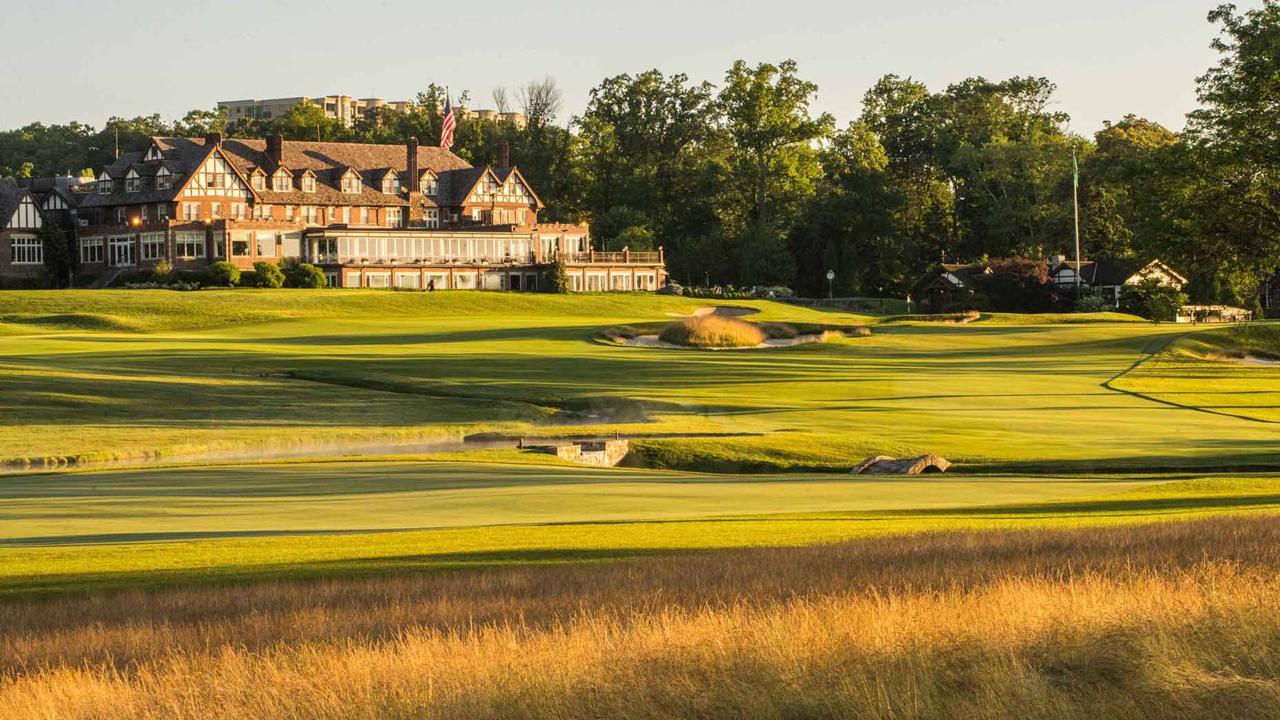 A large clubhouse with Tudor-style architecture overlooks a lush, green golf course—one of the premier PGA Championship venues—surrounded by trees and golden grasses on a sunny day.