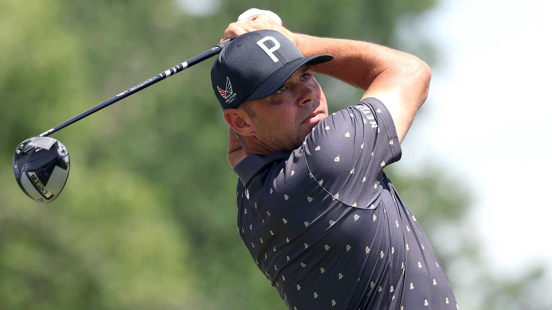 A golfer wearing a dark cap and patterned shirt follows through on a swing during the Charles Schwab Challenge, holding a driver, with a blurred green background of trees.