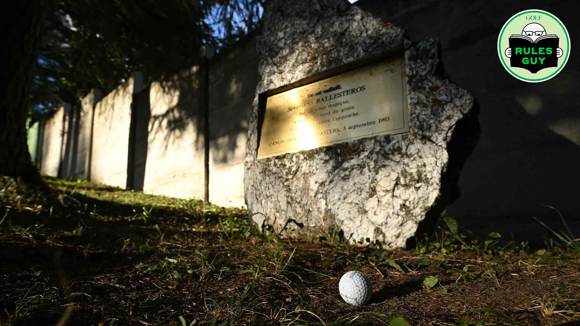 A golf ball rests on grass near a stone memorial plaque embedded in a large rock, sunlight casting shadows from nearby trees—could relief be granted here? A Rules Guy golf logo appears in the top-right corner.