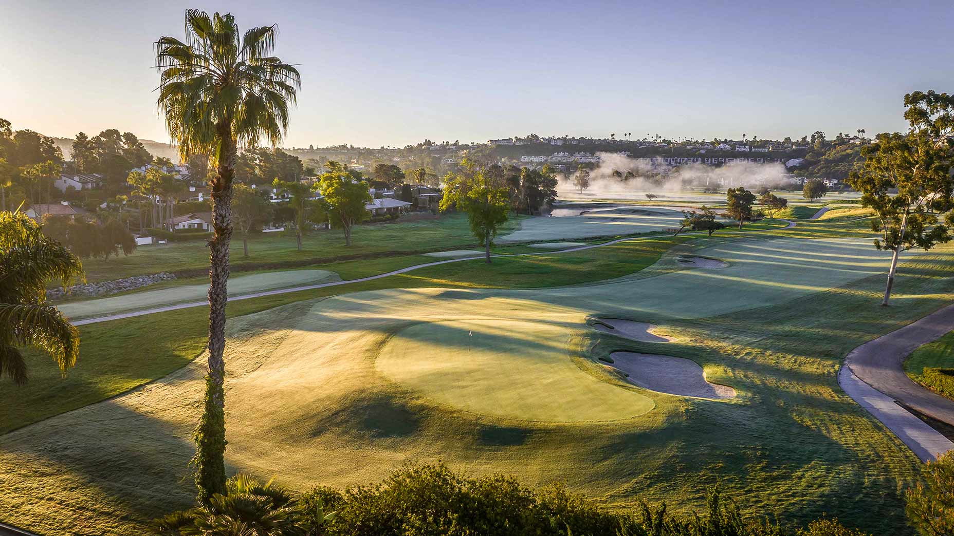 A scenic view of the Omni La Costa golf course at sunrise, with neatly manicured greens, sand bunkers, and a tall palm tree in the foreground; mist rises over a distant water feature, and houses line the horizon.