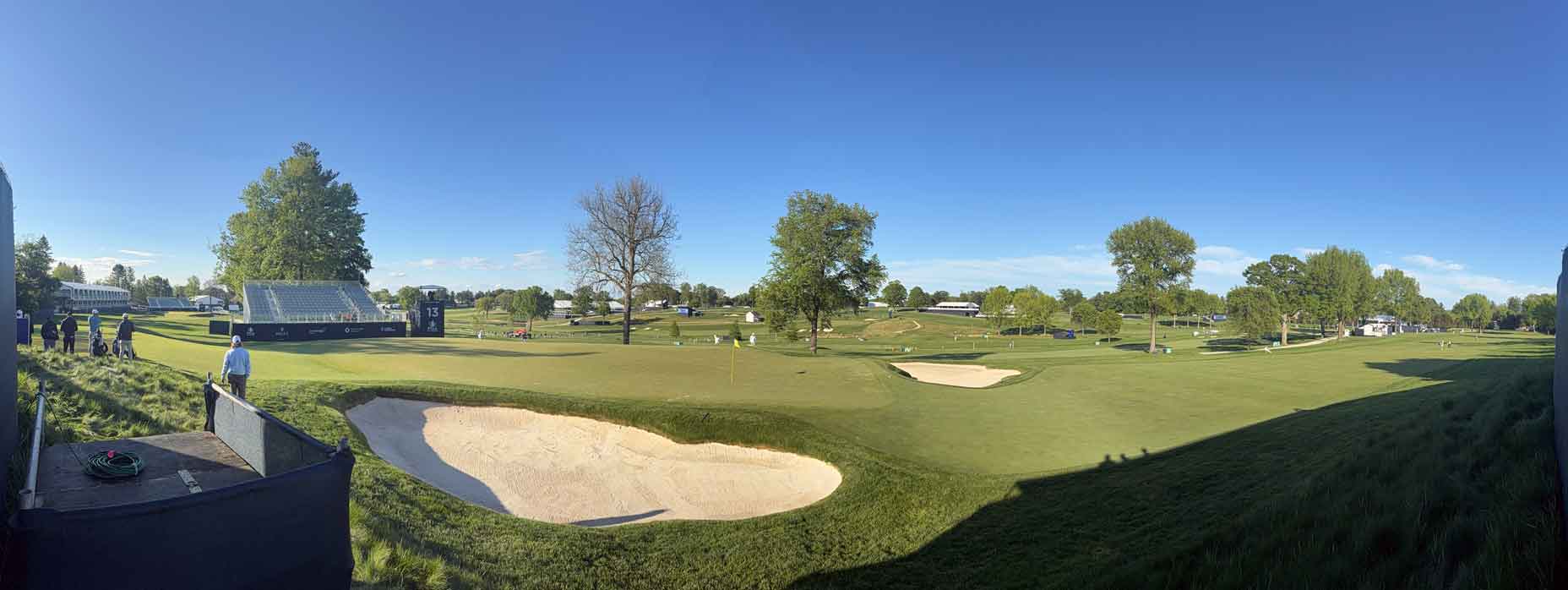 A panoramic view of Aronimink golf course under a clear blue sky, featuring green fairways, sand bunkers, scattered trees, and a few people and structures in the distance—ideal for early observations ahead of the PGA Championship.