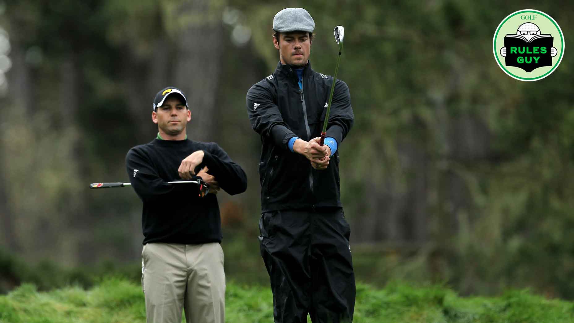 Two male golf partners stand on a grassy course, one in black and khaki adjusting his club, the other in dark rain gear and a flat cap preparing to swing. A Rules Guy golf logo is in the upper right corner, highlighting golf rules.