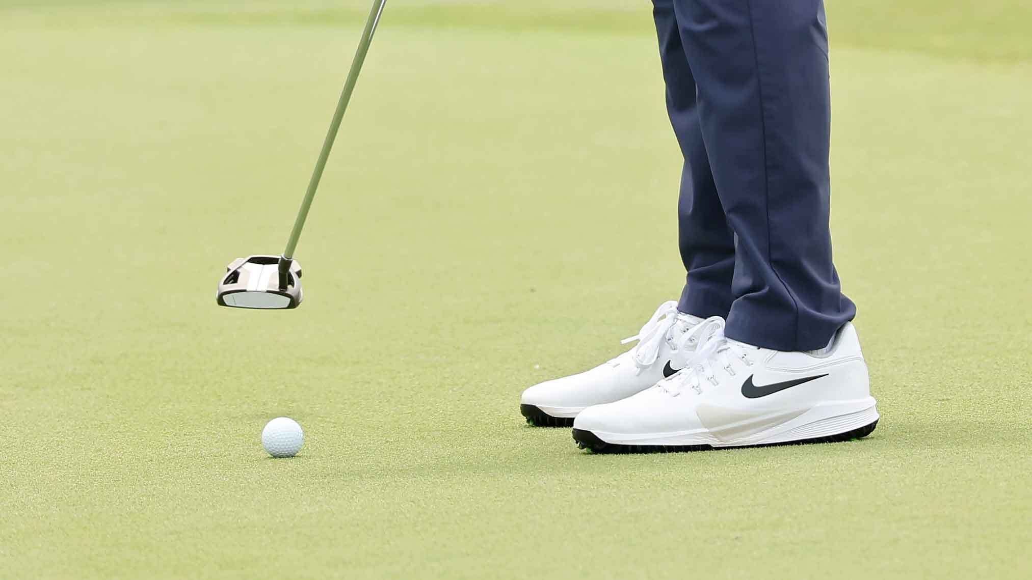 A golfer in white Nike shoes and blue pants demonstrates precise putting technique, positioning the club near the ball on the green for optimal distance control.