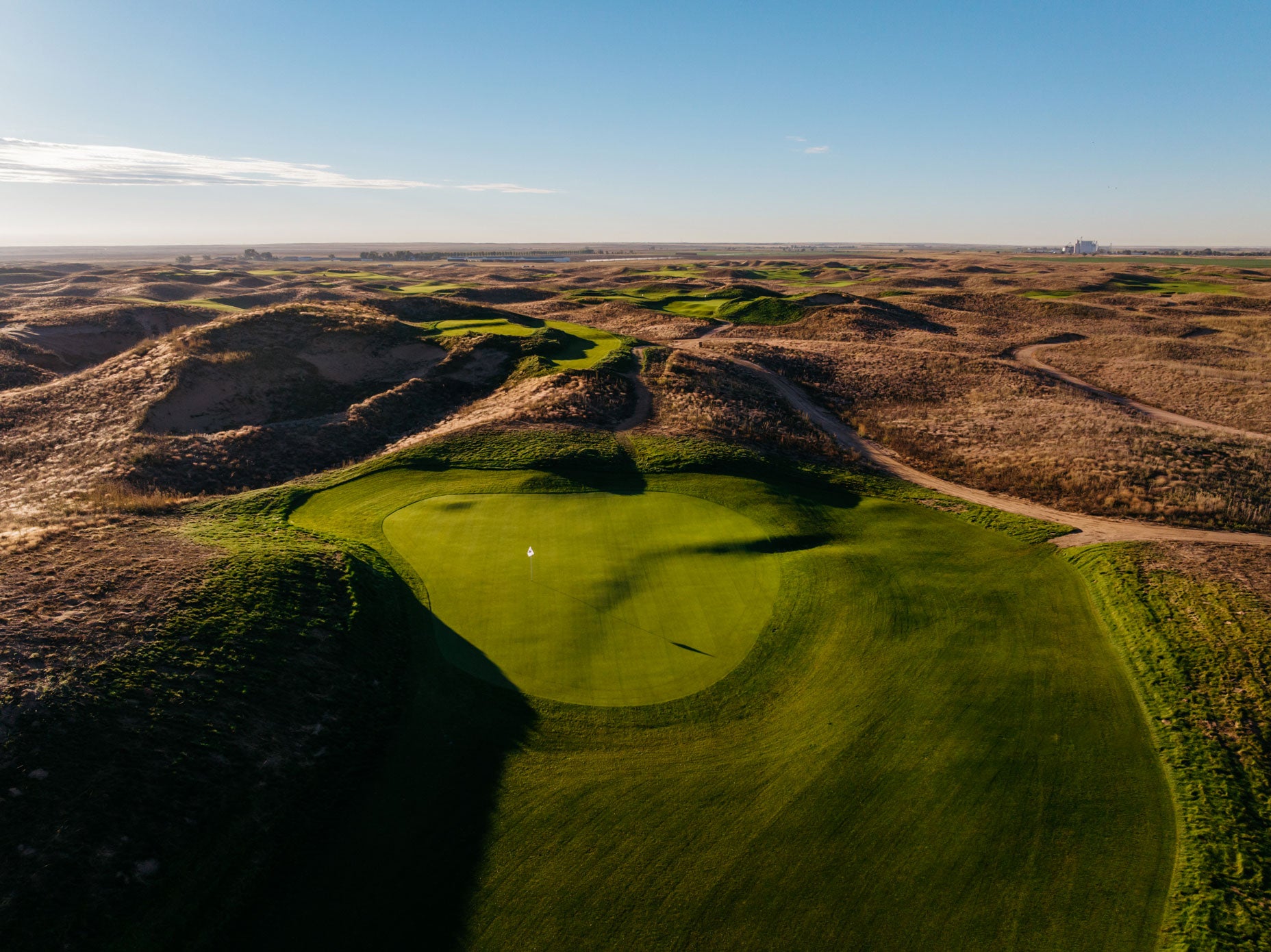 Aerial view of Rodeo Dunes golf course green, sunlit and remote, surrounded by rolling brown grassy hills under a clear blue sky. A flag marks the hole at the center of the green.