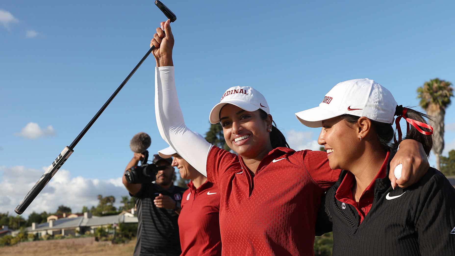 Stanford women golfers, smiling and celebrating on the course after dominating at the NCAA championship. One raises her club triumphantly while the other smiles with an arm around her, as a cameraman films them in the background.