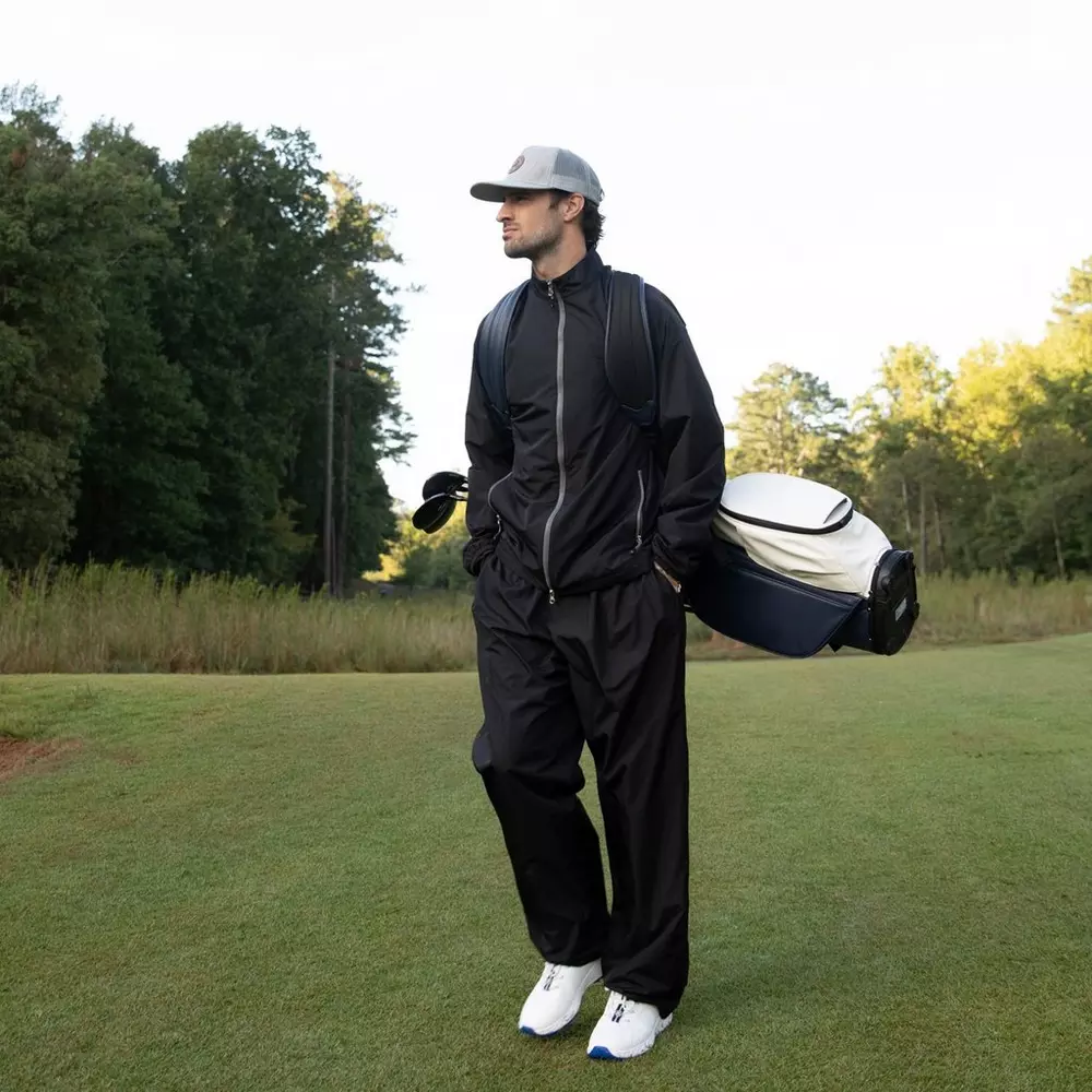 A person wearing a black tracksuit, white sneakers, and a gray cap stands on a grassy field holding a golf bag&mdash;perhaps inspired by the 6 Golf Deals Worth Buying Right Now&mdash;with trees and blue sky in the background.
