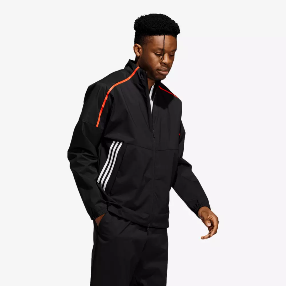 A man wearing a black sports jacket with red shoulder stripes and three white stripes on the side stands with his hands in his pockets, looking down&mdash;perfect attire for casually browsing 6 Golf Deals Worth Buying Right Now. The background is plain and light-colored.