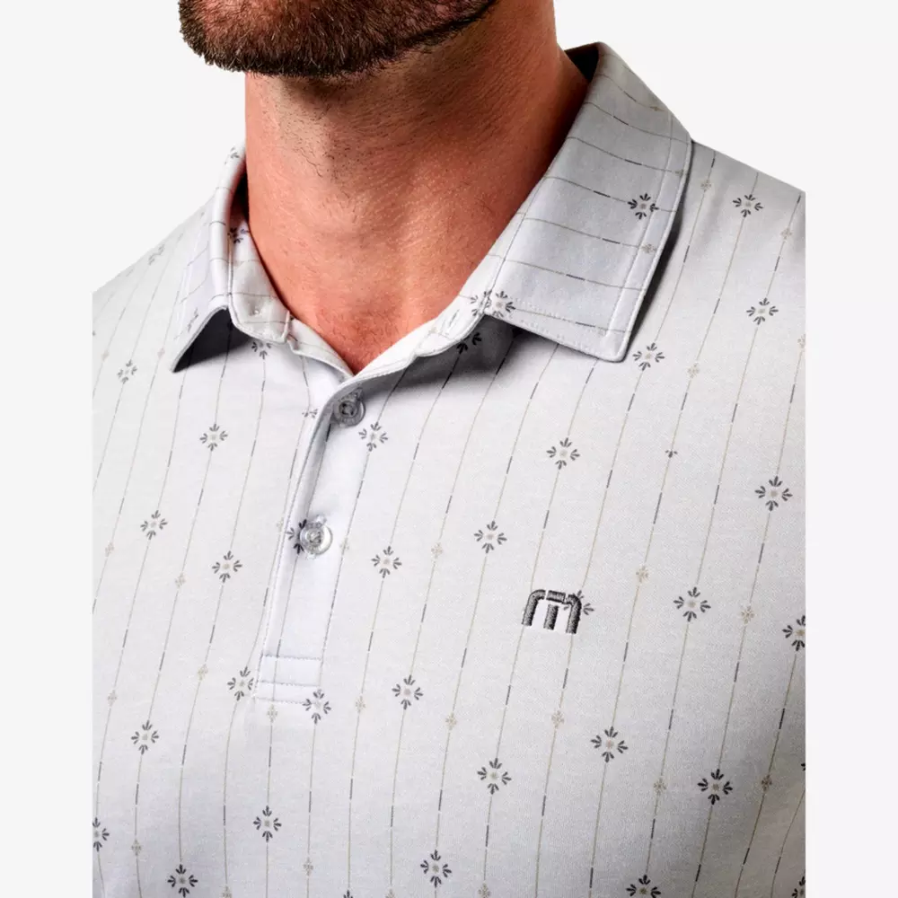 A close-up of a man wearing a light gray, short-sleeve polo shirt with a subtle, dark geometric pattern and an embroidered logo on the chest. The shirt has a collar, two buttons, and he&rsquo;s also sporting the best golf smartwatch on his wrist.