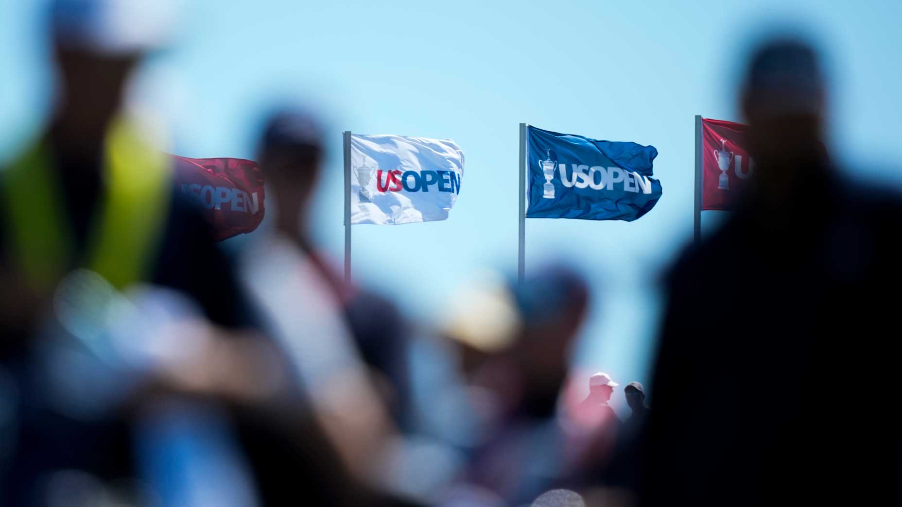 U.S. Open flags wave in the brutal winds against a blue sky, with blurred silhouettes of people in the foreground, at Shinnecock Hills.