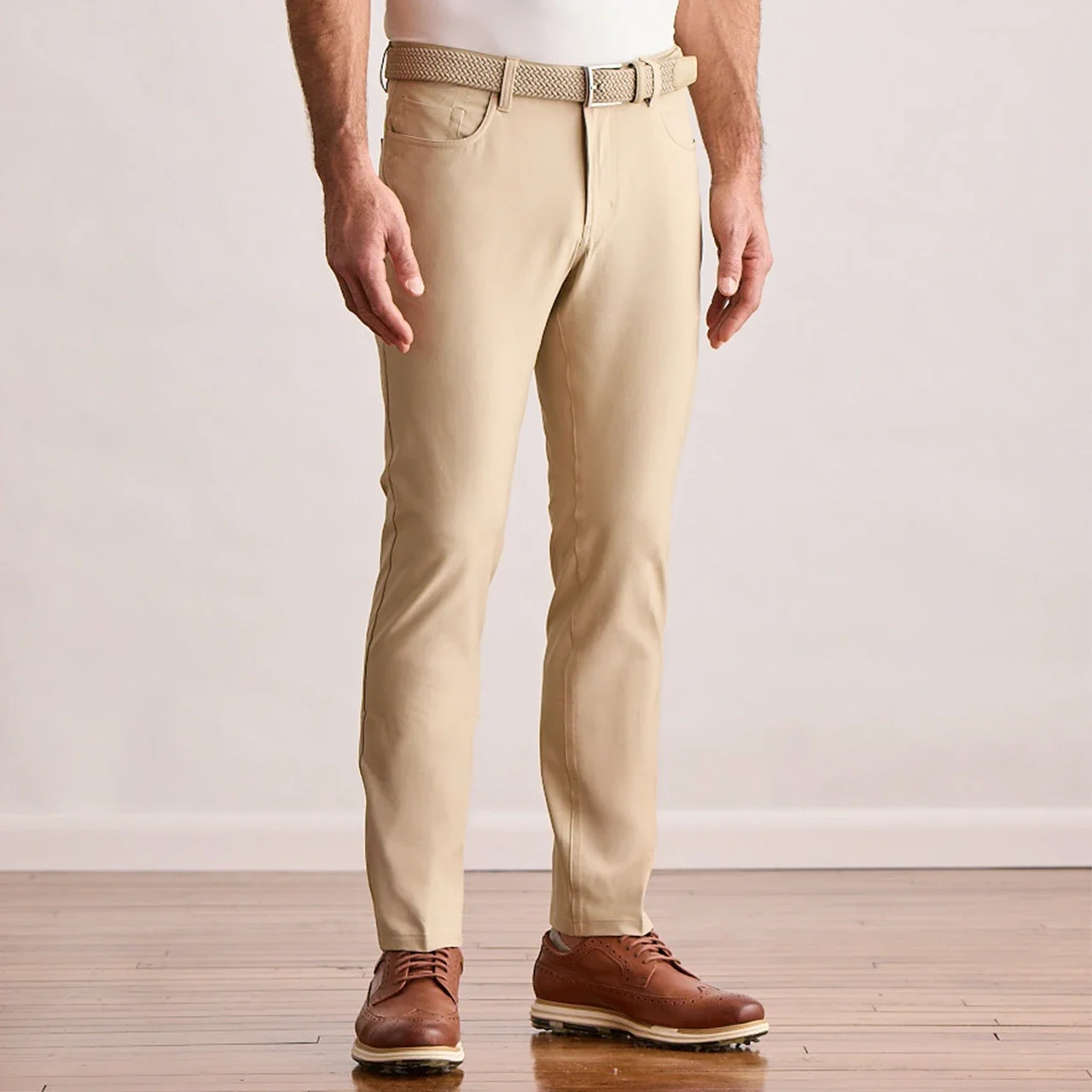 A man wearing beige slim-fit pants from a Top 5 apparel brand, paired with a woven belt and brown leather shoes, stands on a wooden floor against a plain light-colored wall.