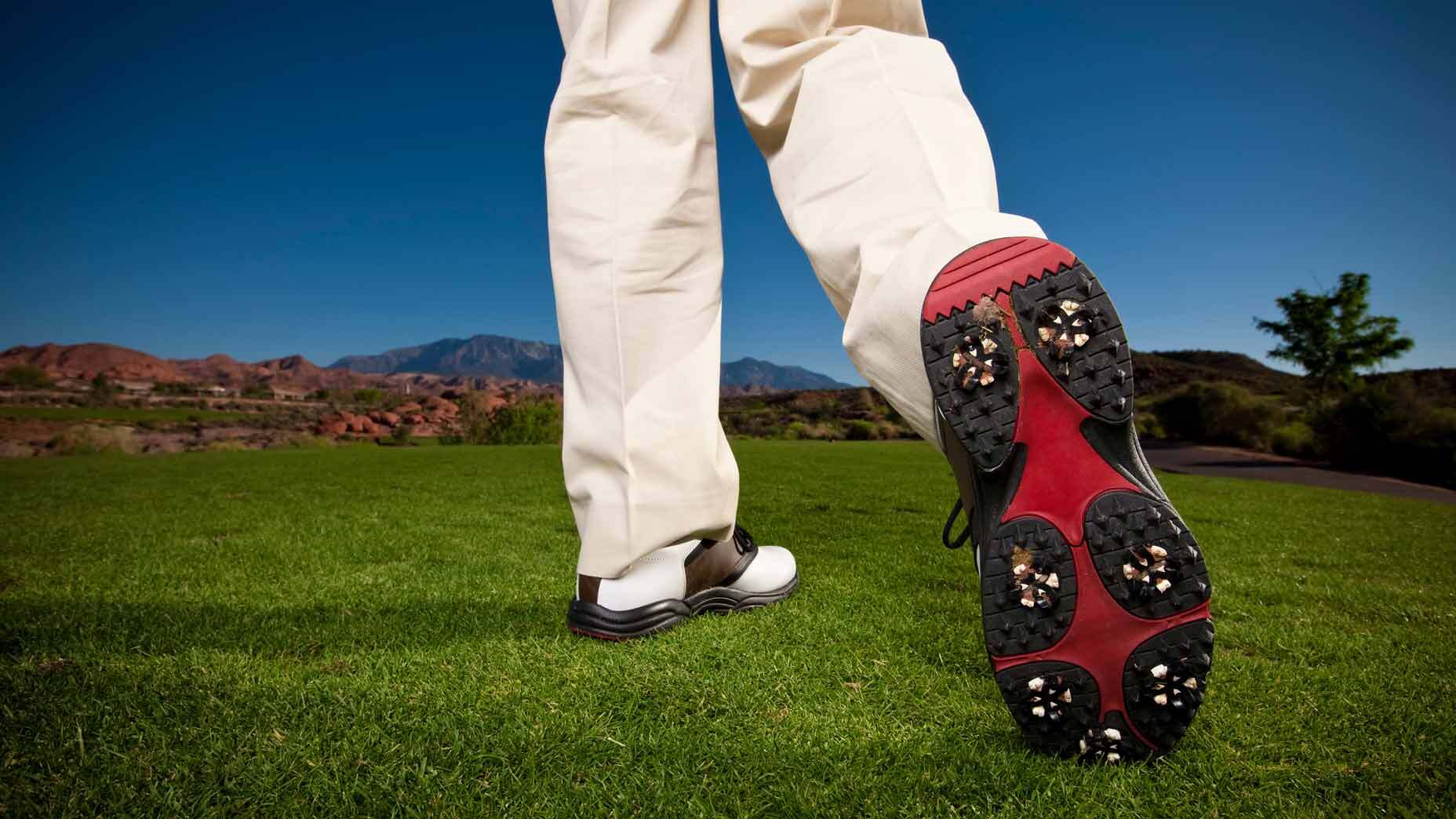 A golfer stands on green grass, showing the spiked sole of one Arccos golf shoe, with mountains and a clear blue sky in the background.