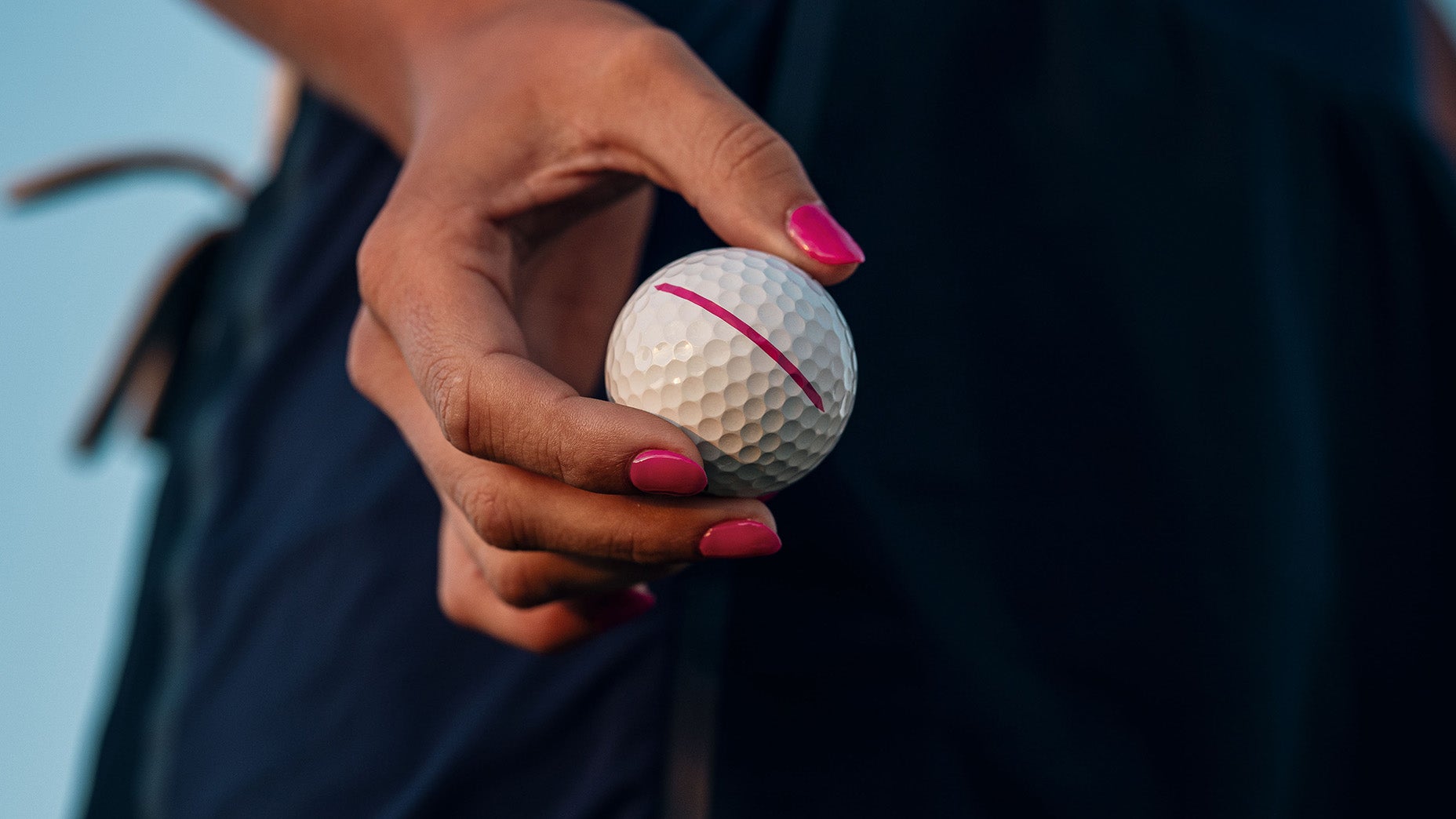A person with pink-painted nails holds a white golf ball marked with a pink line, against a blurred dark background.