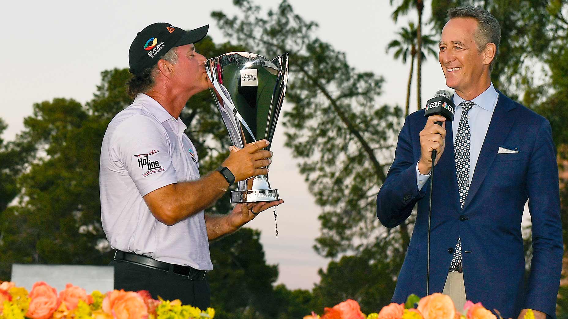 A golfer kisses a large silver trophy while Rich Lerner, in a suit, holds a microphone and smiles. They are standing outdoors, with flowers in the foreground and trees silhouetted against the sunset.