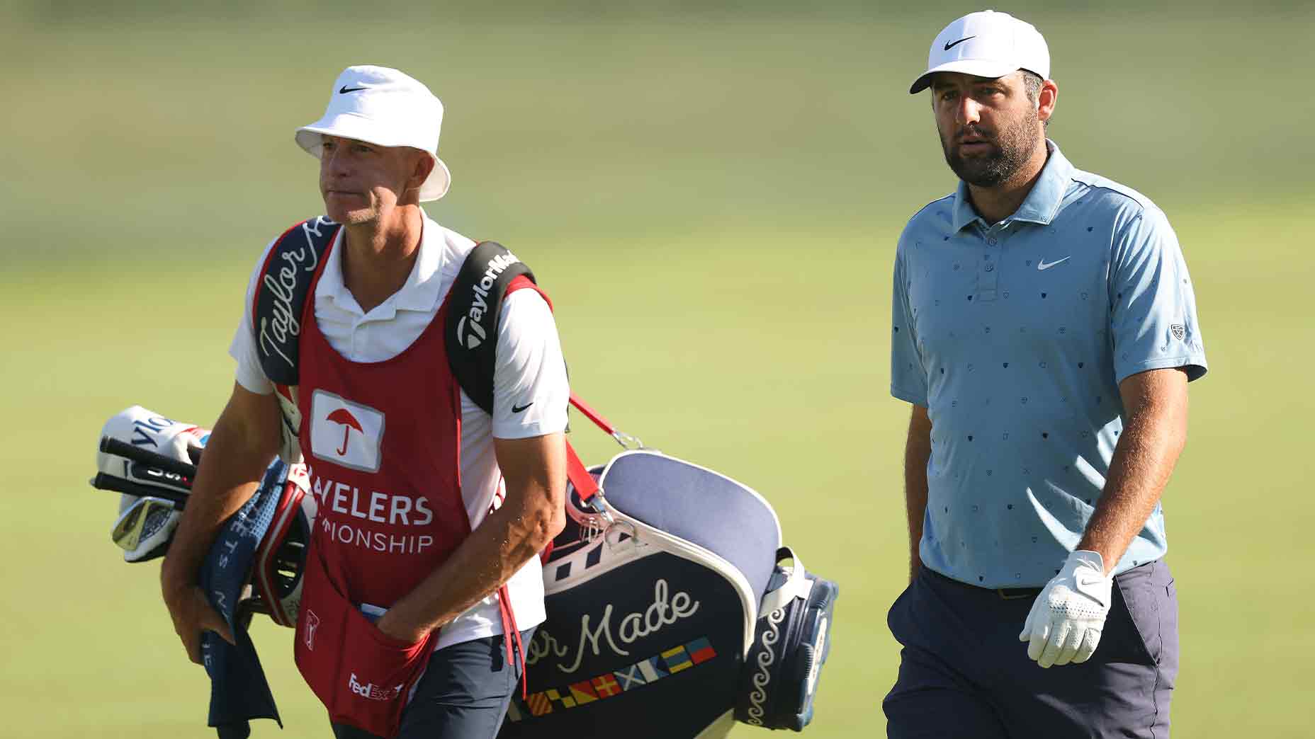 Two men walk on a golf course during the 2026 Travelers Championship; one wears a white hat and red caddie vest carrying golf clubs, while the other sports a blue polo shirt, white cap, and glove holding a golf tee.
