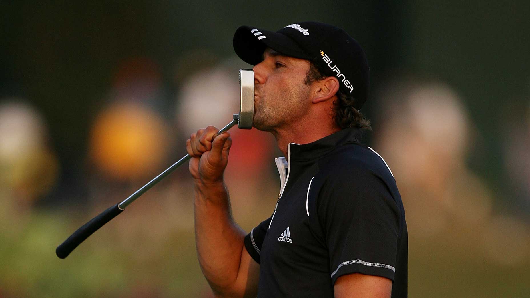 A golfer wearing a black Adidas shirt and cap kisses his golf putter, celebrating on a golf course with a blurred crowd in the background.