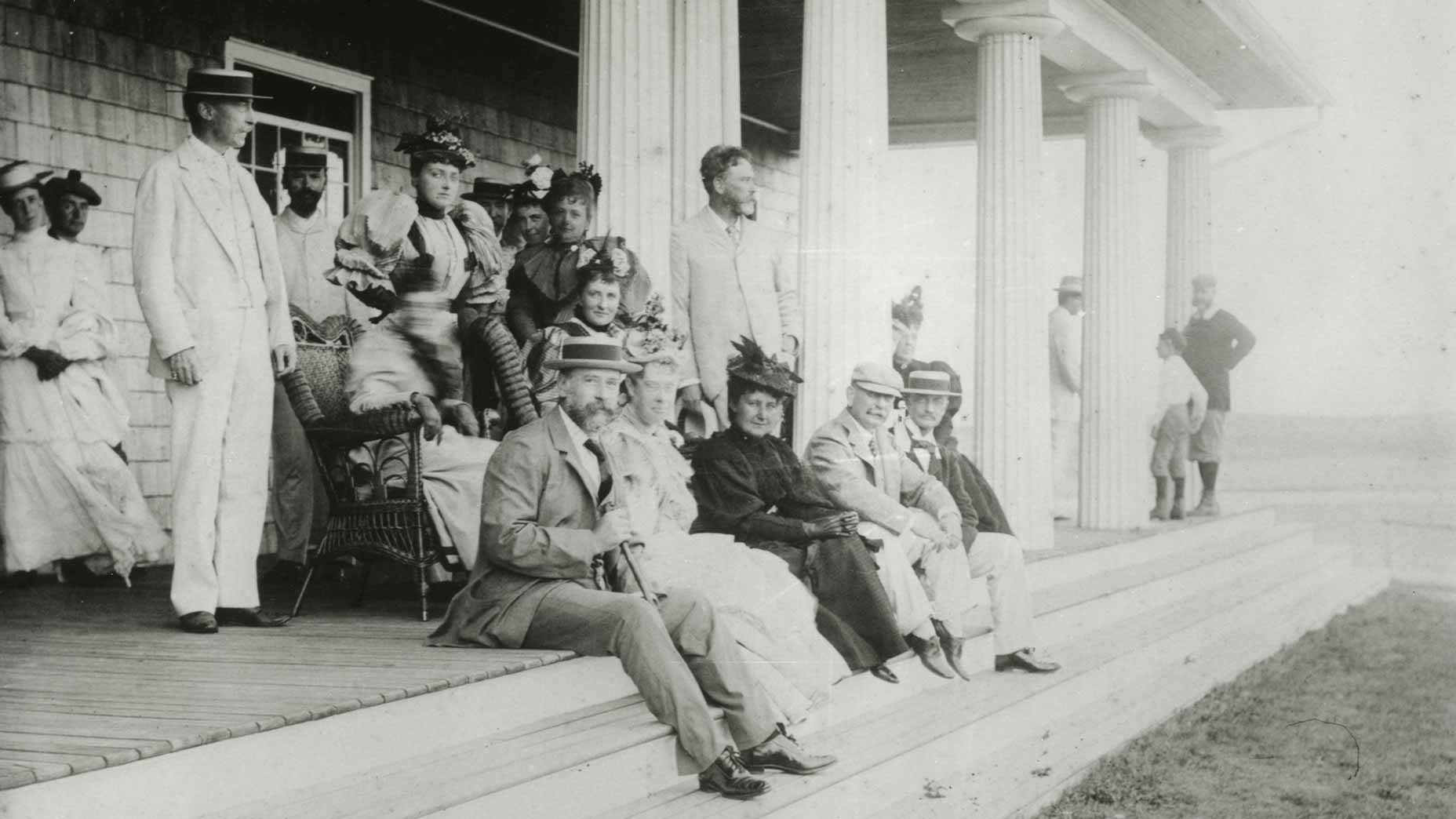 A group of men and women in formal late 19th-century attire gather on the steps of a columned building, posing for a photograph. The relaxed mood suggests they may be attendees of the 1896 U.S. Open at Shinnecock Hills.