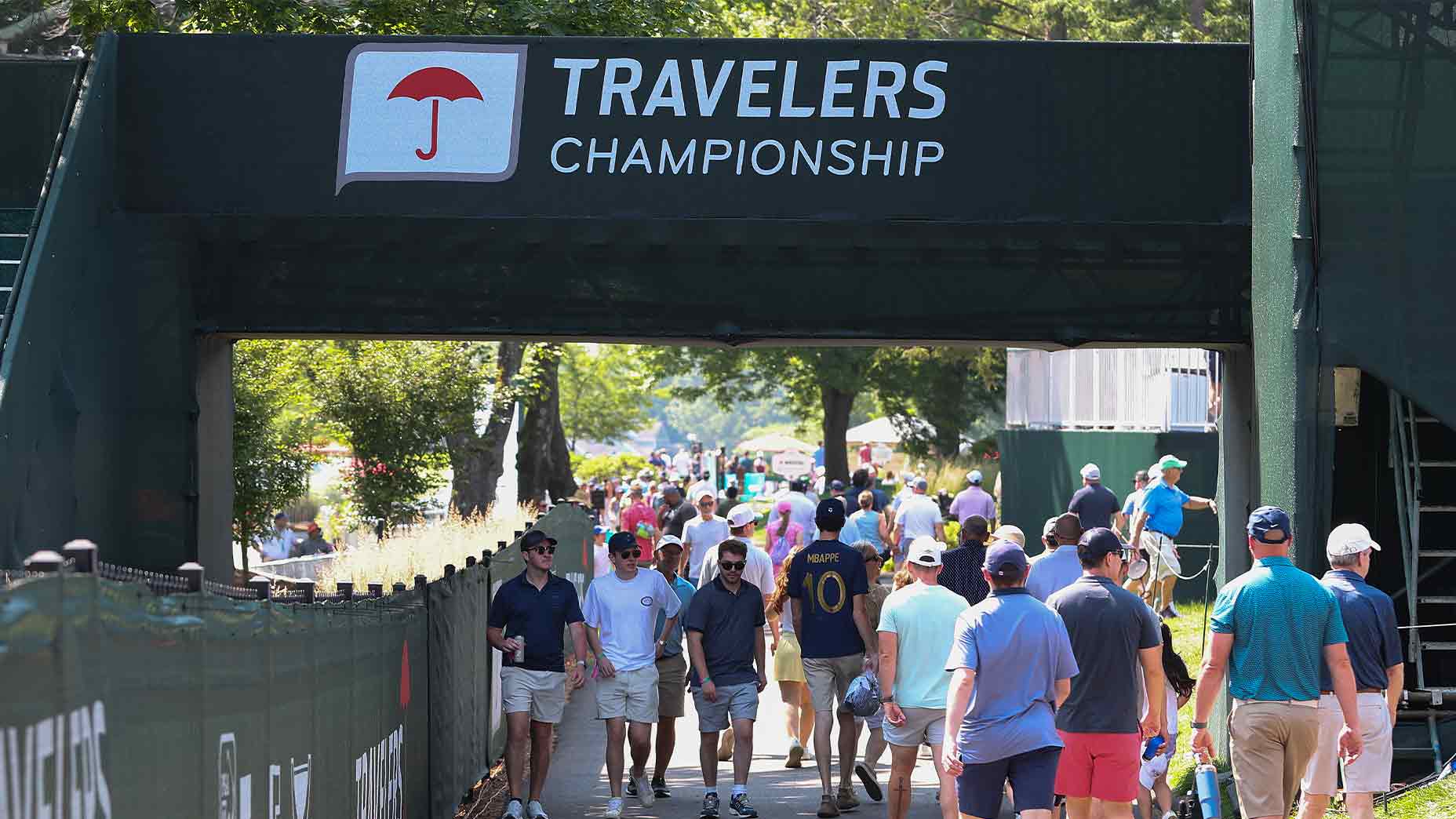 A crowd of people walk under a large sign reading 2026 Travelers Championship at a golf event, with trees and fencing visible on a sunny day.