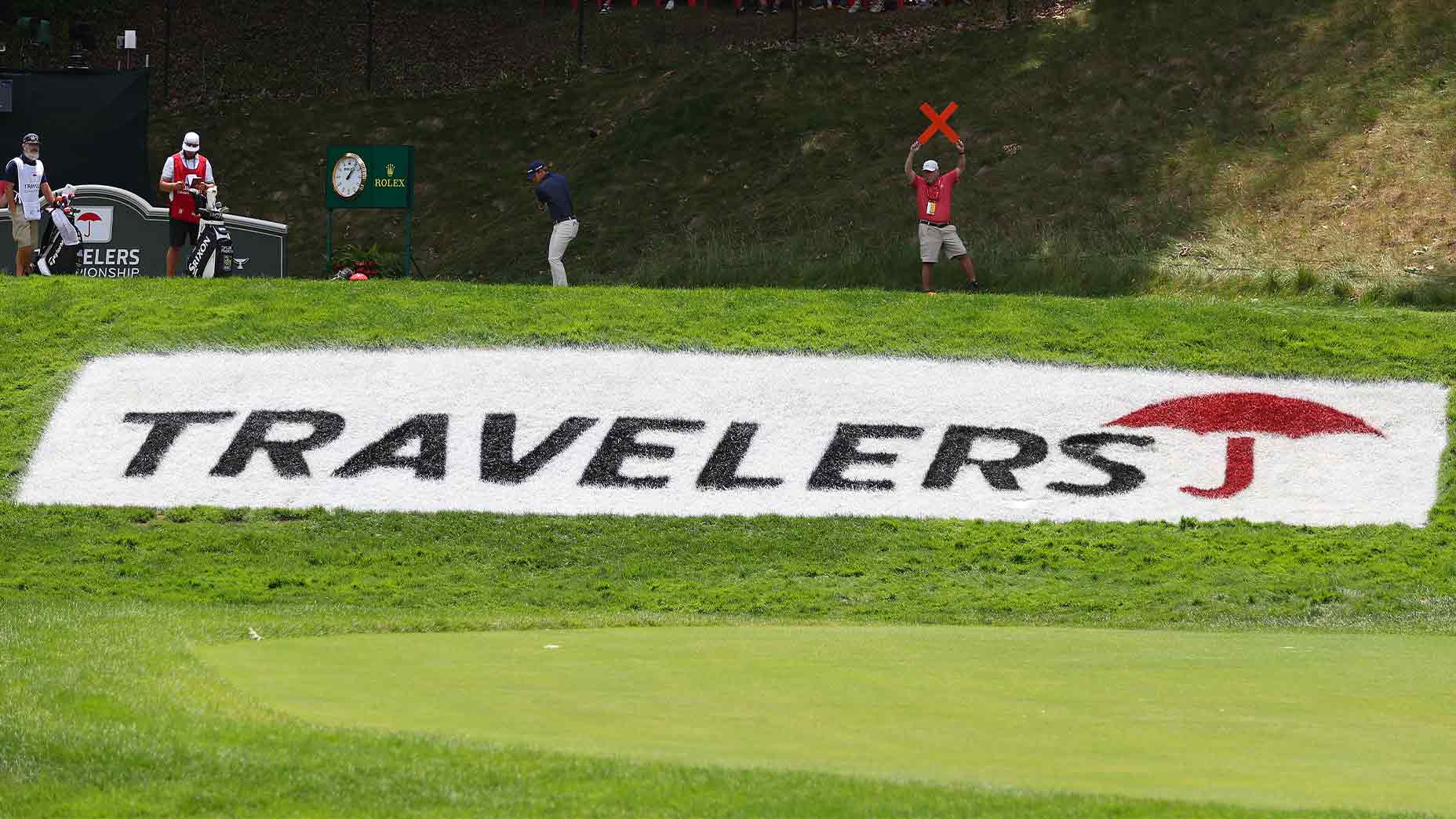 A golf course with a large white TRAVELERS sign and red umbrella logo, as a golf official signals with a red X and players stand nearby—setting the stage for the 2026 Travelers Championship Thursday tee times.