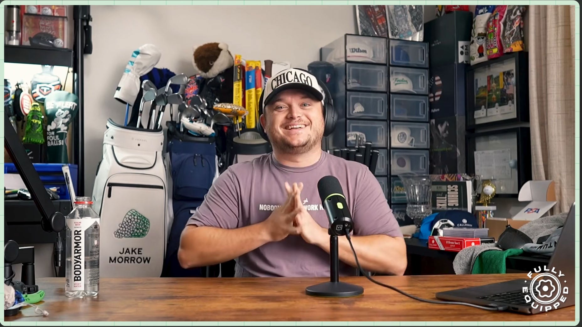 A man wearing a CHICAGO cap and purple t-shirt sits smiling at a desk with a microphone. Behind him are shelves, golf bags, and sports gear. A water bottle and laptop are on the desk.