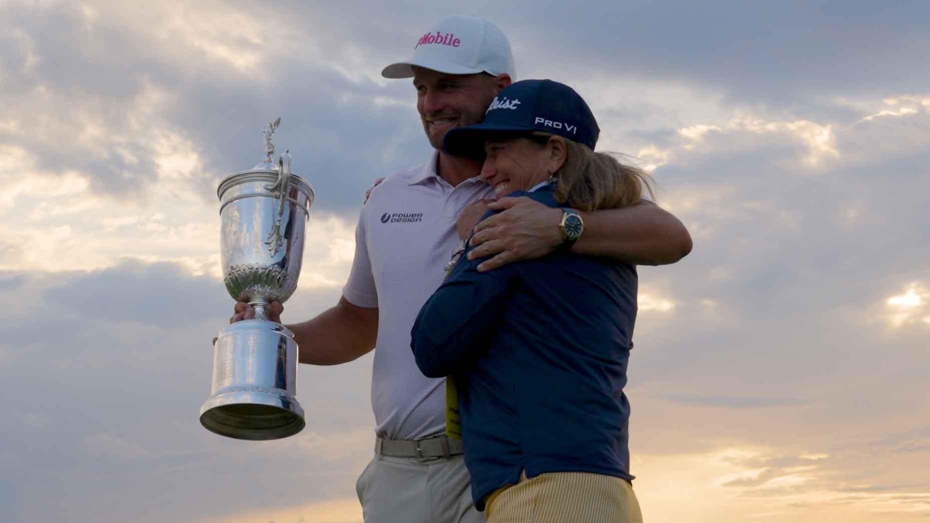 A smiling Wyndham Clark in a white cap holds a silver trophy and hugs a woman in a blue cap and jacket, both standing outdoors with a cloudy sky and soft sunlight in the background.