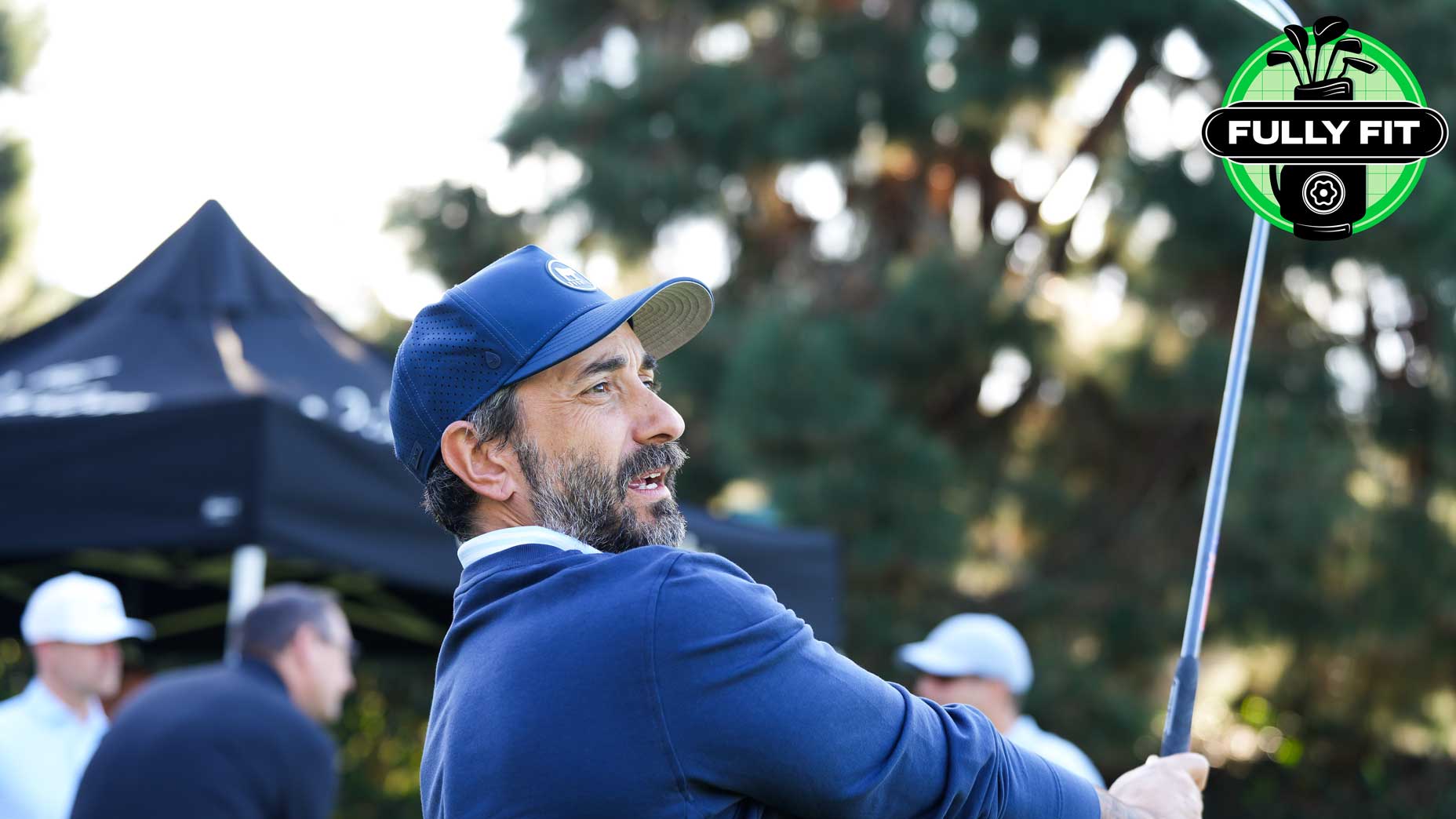 A man in a navy jacket and cap swings a golf club outdoors, showcasing his golf game, with trees and a black canopy tent in the background. Several people are visible behind him. A Fully Fit logo appears in the top right corner.