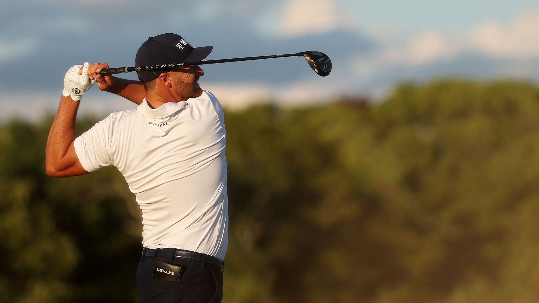 A golfer in a white shirt and dark cap follows through on his swing, holding a golf club on a sunny day with green trees and a blue sky in the background.