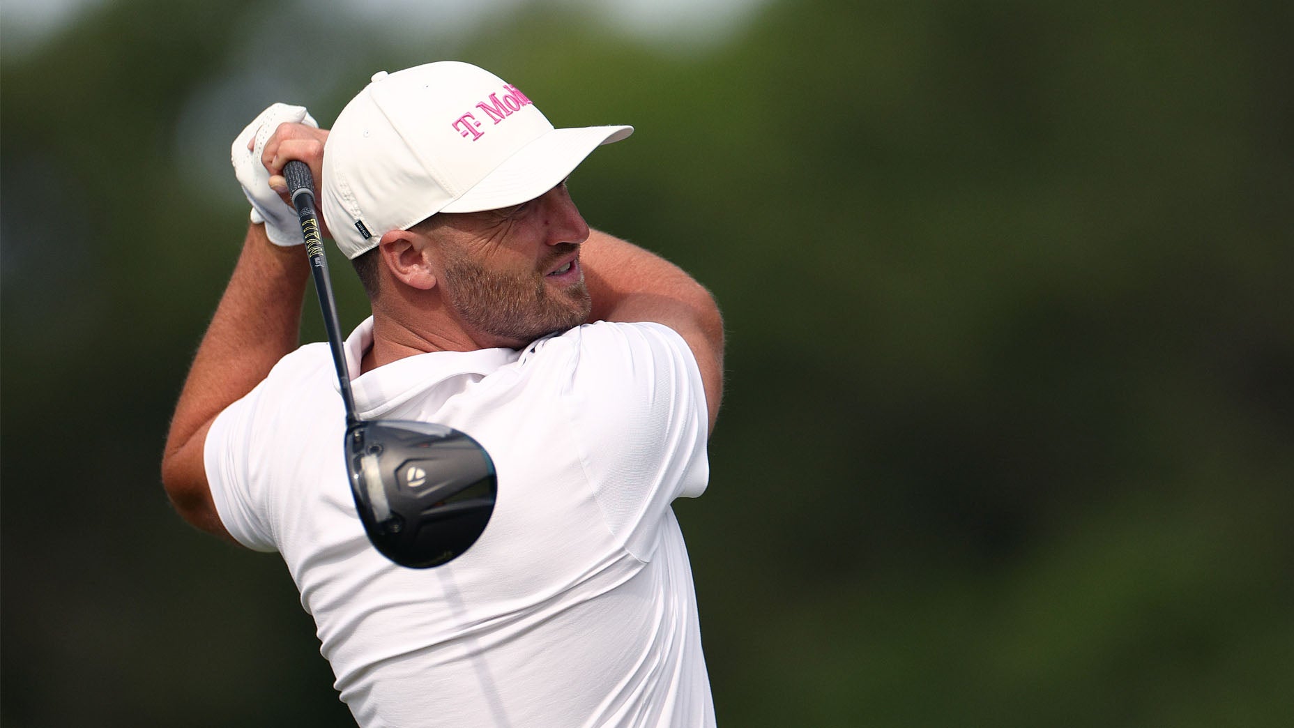 A golfer wearing a white shirt and cap swings a driver club on a golf course, with a blurred green background.