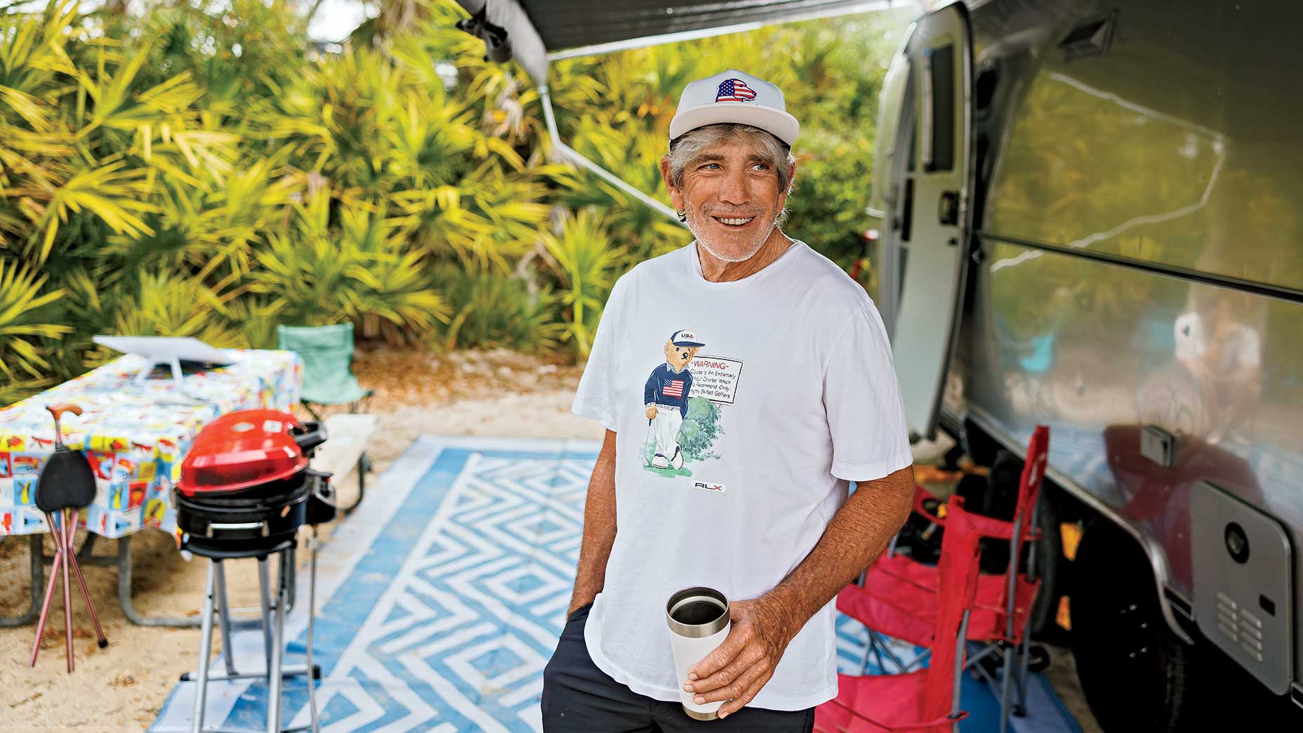 A smiling older man, a peculiar golf coach who lives the dream, stands by his RV in a graphic tee and cap, holding a cup. A red grill, colorful tablecloth, chairs, tropical plants, and a blue outdoor mat complete the scene.