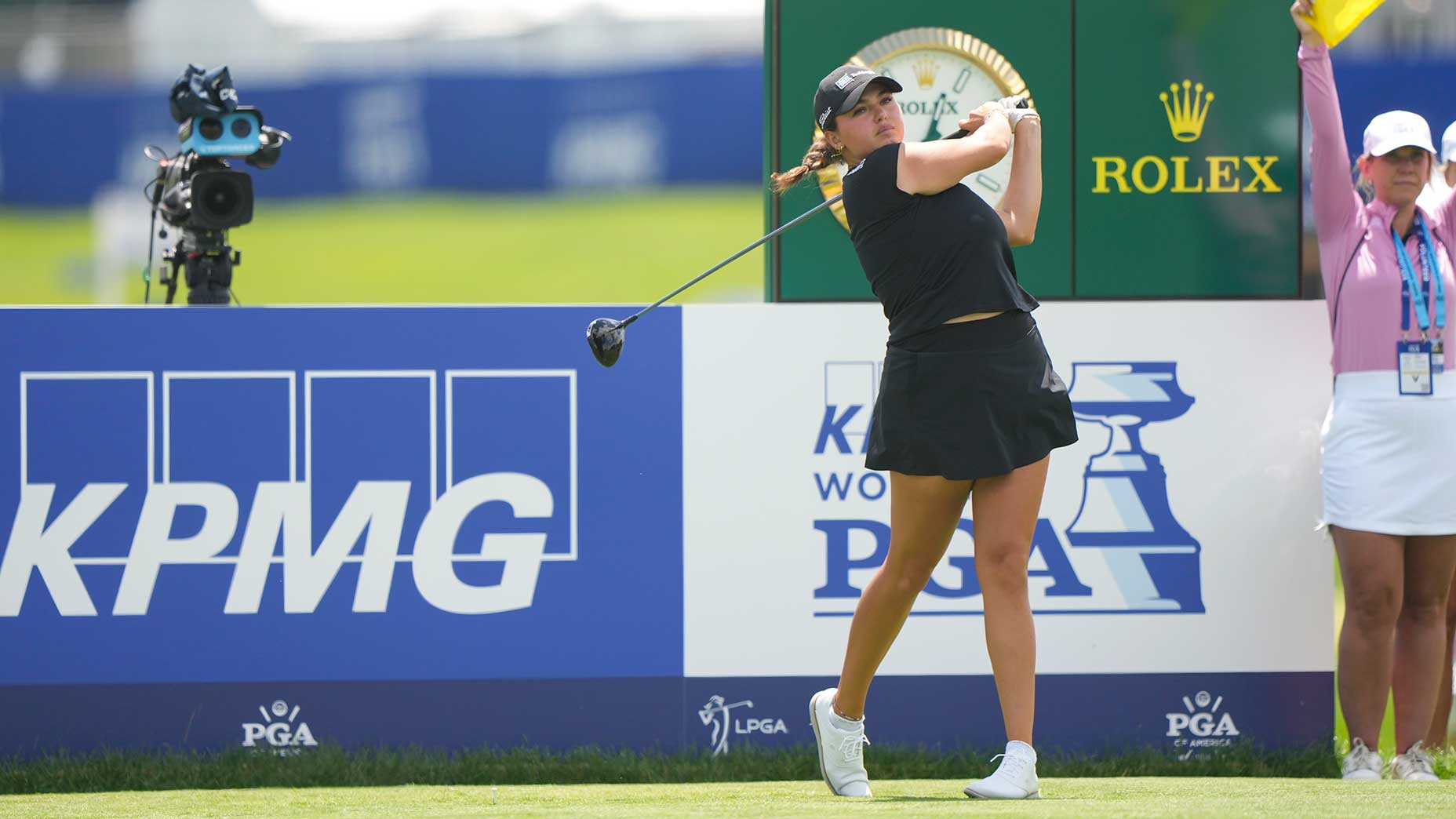 A female golfer in a black outfit watches her drive after swinging, with a camera, Rolex clock, and KPMG and PGA signage visible in the background. An official in pink stands nearby holding a green flag.