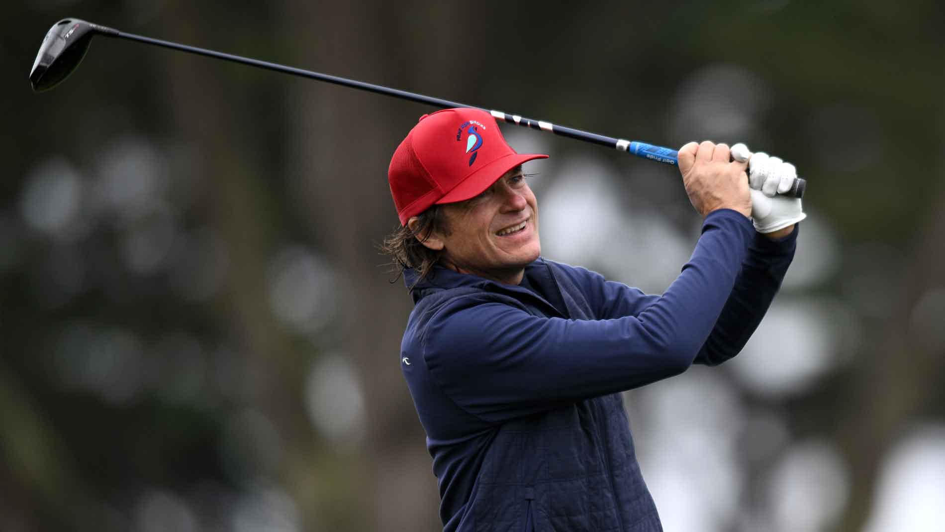 jason bateman wearing a red cap, navy vest, and white glove is smiling while swinging a golf club outdoors at Bel-Air, with blurred trees in the background.