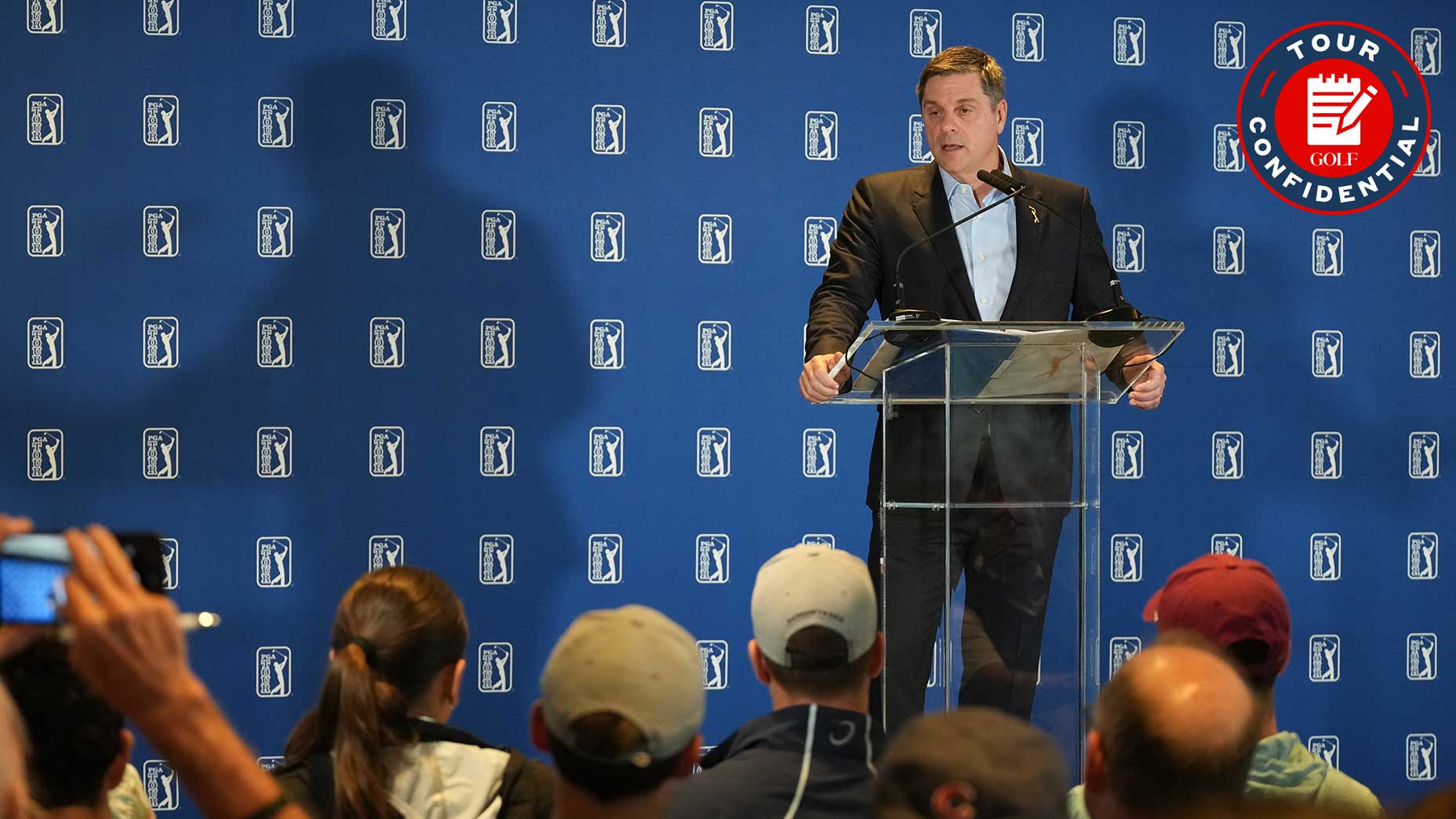 A man in a suit speaks at a clear podium in front of a PGA Tour backdrop, addressing an audience. The Tour Confidential logo appears in the top right corner.