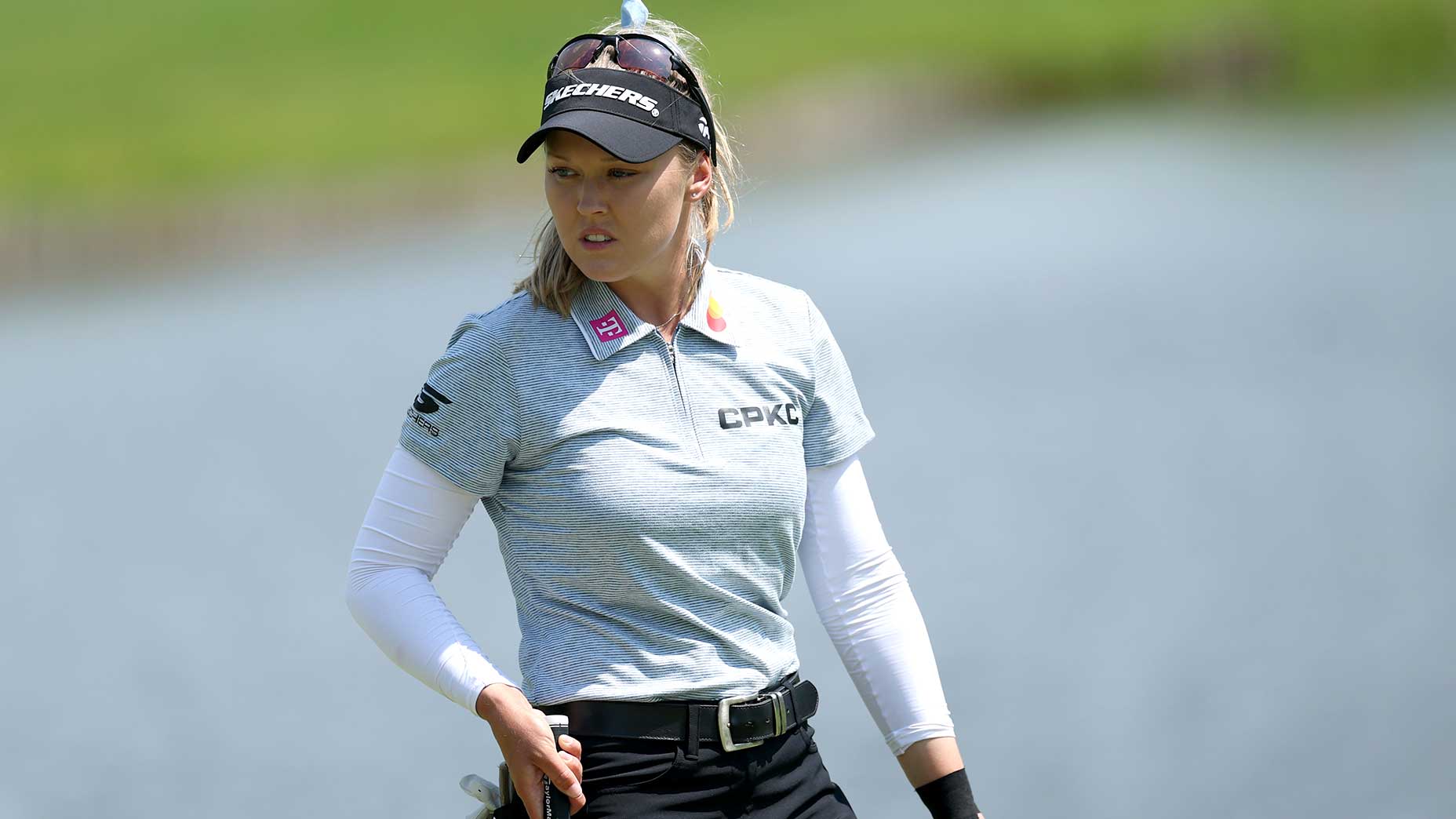 A female golfer wearing a cap and sunglasses stands on a golf course, holding a putter. She is dressed in a light grey shirt with white sleeves and black pants, looking focused with water visible in the background.