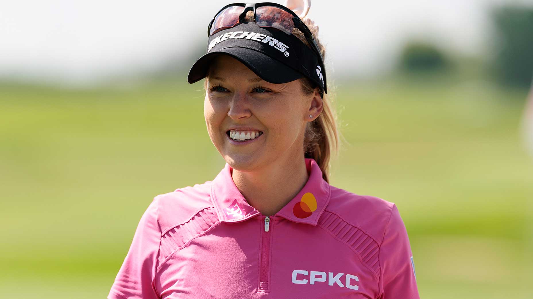 A smiling female golfer in a pink shirt and black visor stands outdoors on a golf course, with sunglasses resting on her visor. She is ready for the KPMG Women's PGA Championship—don’t miss the featured groups to see how to watch her in action!.