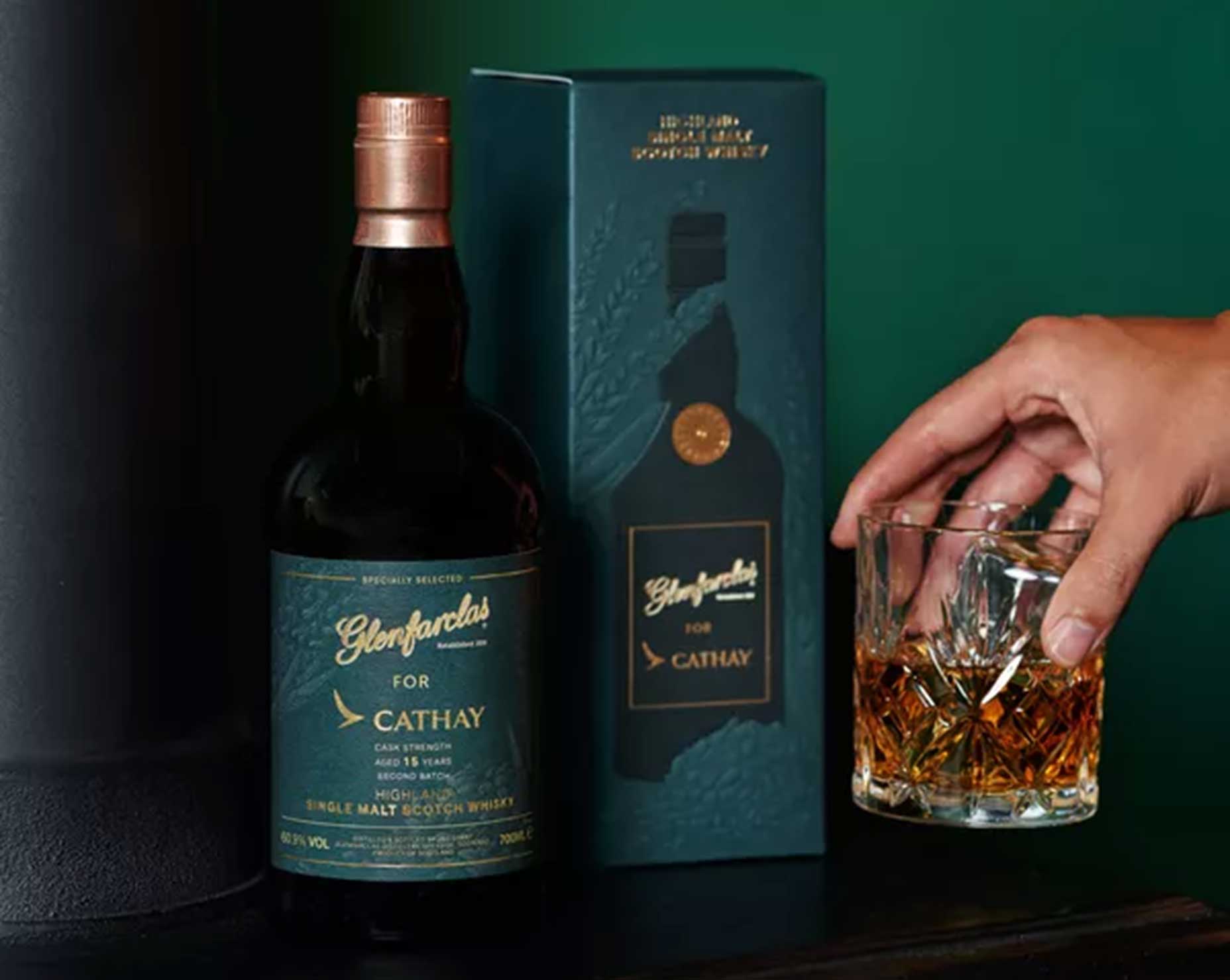 A hand holds a glass of whisky next to a Glenfarclas For Cathay single malt Scotch whisky bottle and its matching dark green box, set against a dark background—an exceptional gift for Father’s Day and fans of fine whiskies.