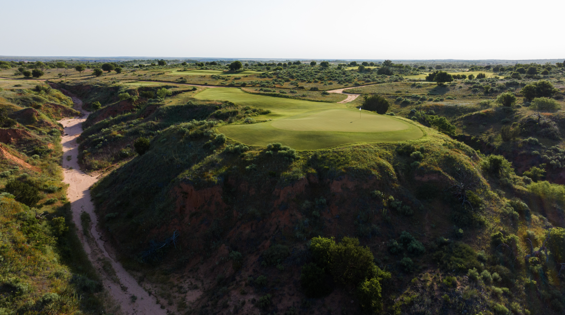 childress hall green golf course sits atop a steep, grassy hill surrounded by rugged terrain and winding dirt paths—one of the hottest hangouts for golf lovers. Sparse vegetation and roaming cattle create a natural, scenic setting under a clear sky.