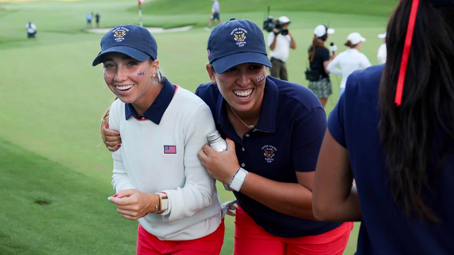 Two women in matching caps and red pants, smiling and walking arm-in-arm on a golf course. Both have flag stickers on their cheeks and appear to be celebrating, with other people and cameras in the background.