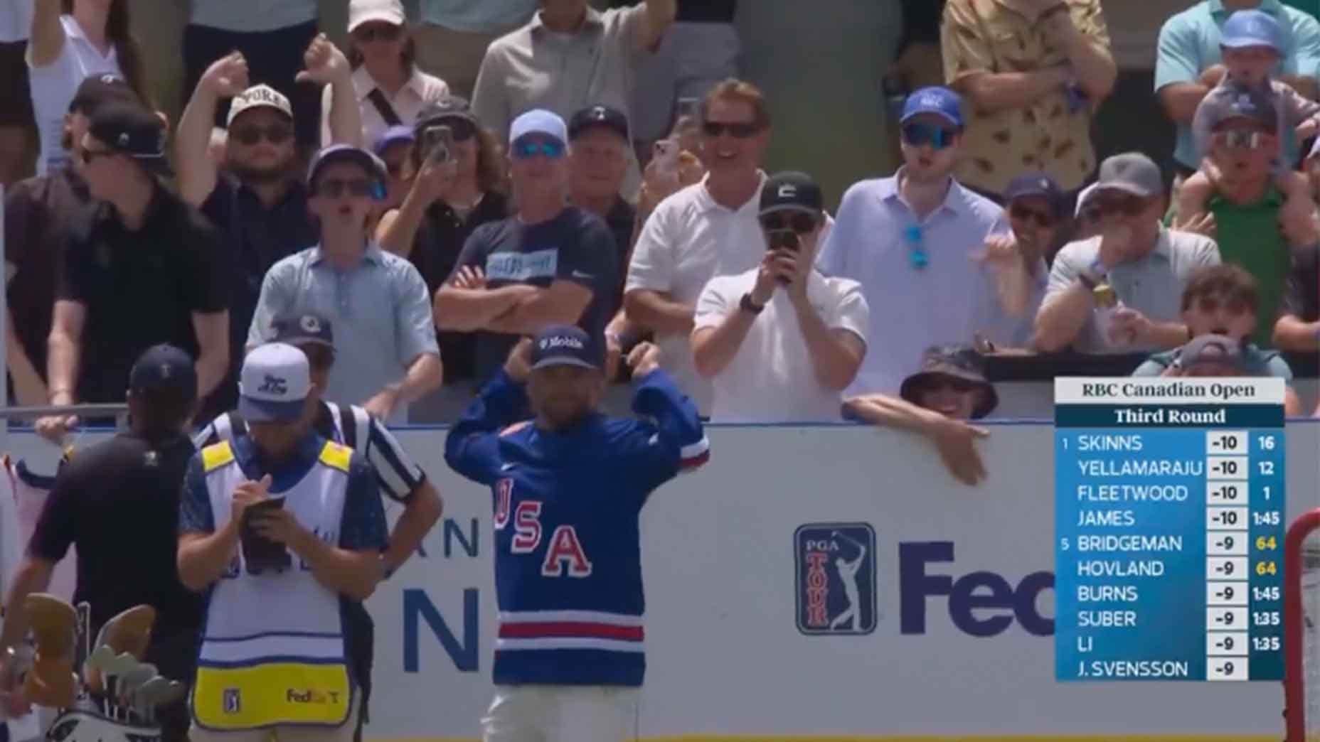 A golfer wearing a blue USA hockey jersey stands with hands on head, looking surprised or disappointed, as spectators behind him react. A leaderboard is shown on the right side of the screen.