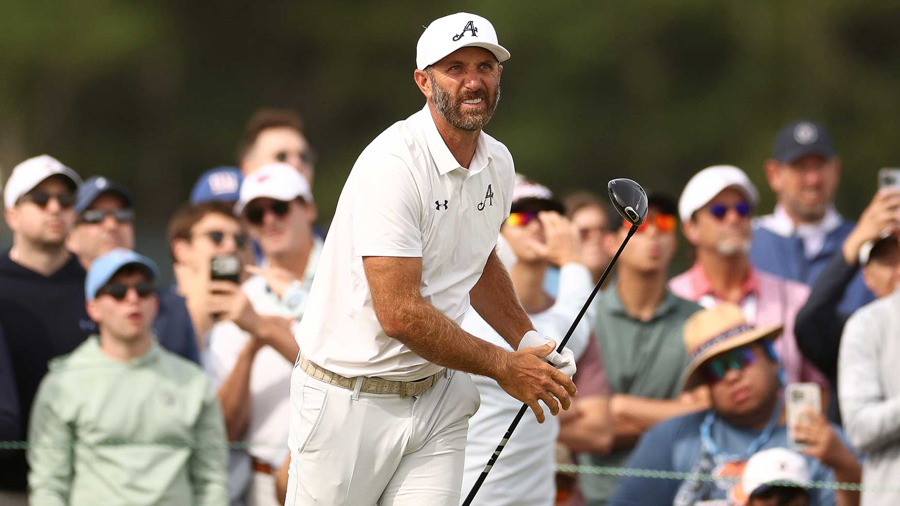 A golfer in a white shirt, pants, and cap holds a golf club after a swing, as the crowd watches—reminiscent of Dustin Johnson facing the pressure of a disastrous Friday on the golf course.