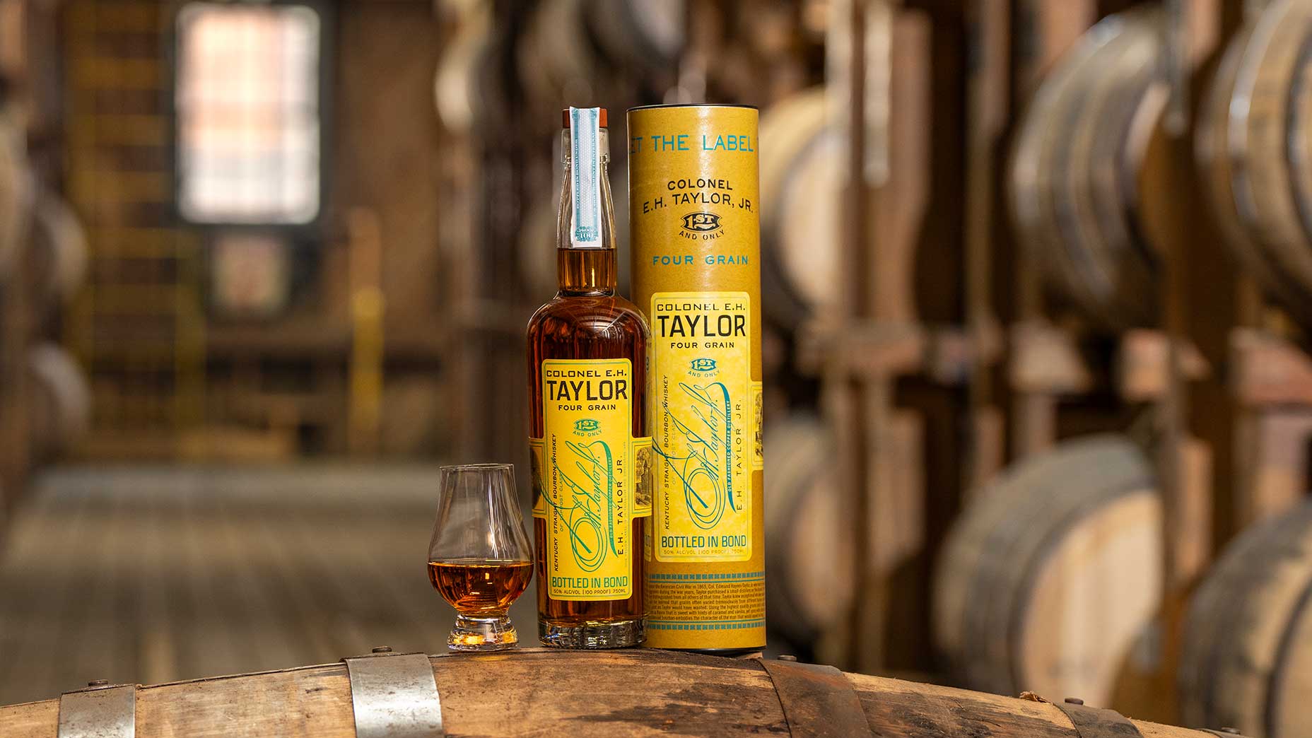 A bottle and glass of Colonel E.H. Taylor Four Grain bourbon sit on a barrel, with the bottle’s yellow box beside them—an exceptional Father’s Day gift. Wooden whiskey barrels line the blurred background inside a distillery warehouse.