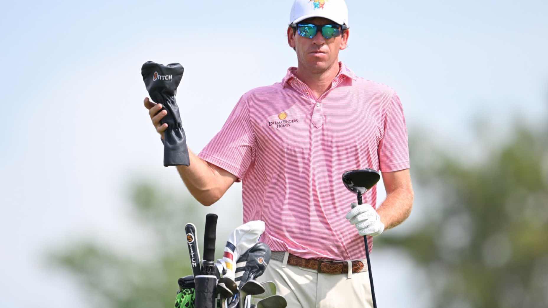 A golfer wearing sunglasses and a pink striped polo shirt stands next to his golf bag, holding a club head cover in one hand and a driver in the other, with a blurred outdoor background.