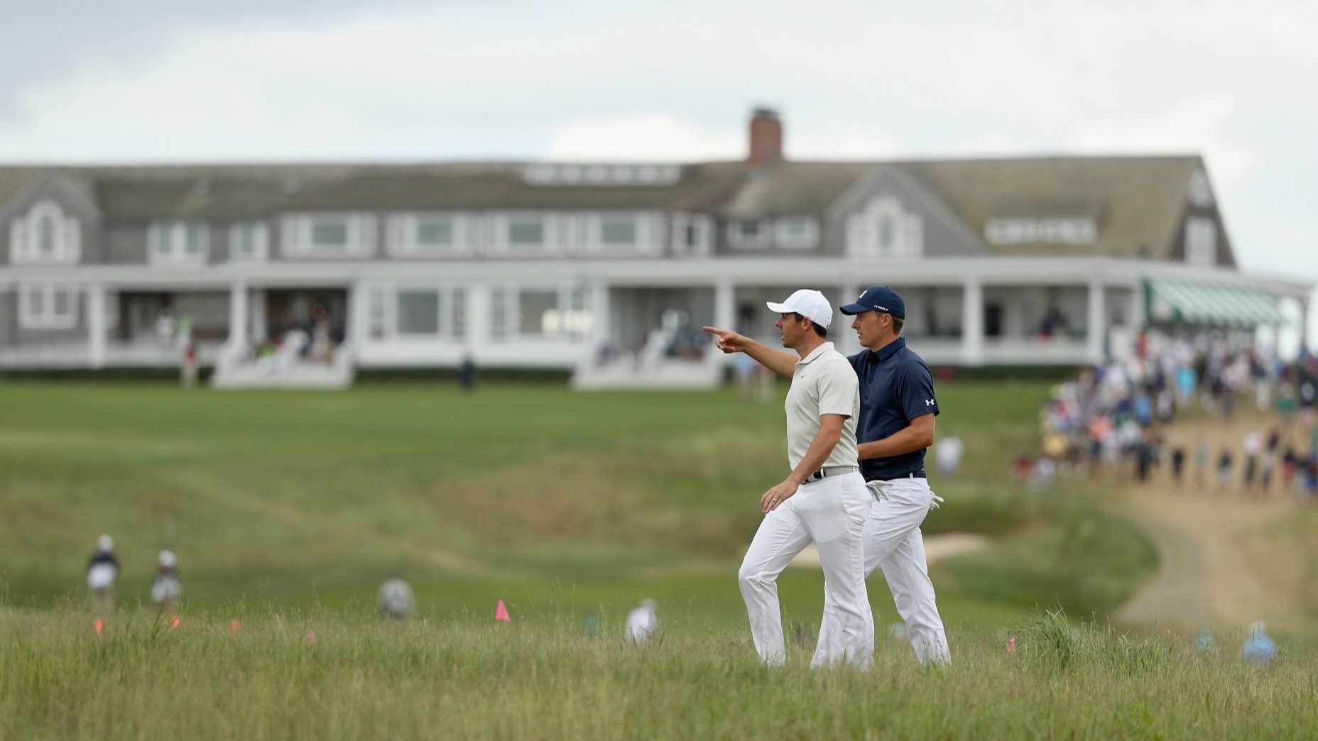 Two golfers, including Rory McIlroy, walk at Shinnecock —one pointing ahead. A large clubhouse with columns and people on its porch is visible in the background, along with spectators watching from the side.