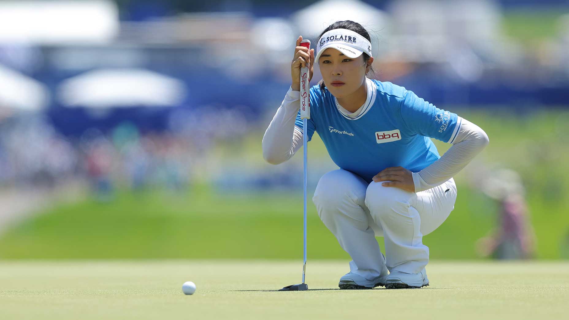 A female golfer in a blue shirt and white pants squats on the green, focusing intently as she lines up her putt with a golf club, with a blurred background of a golf course and spectators.