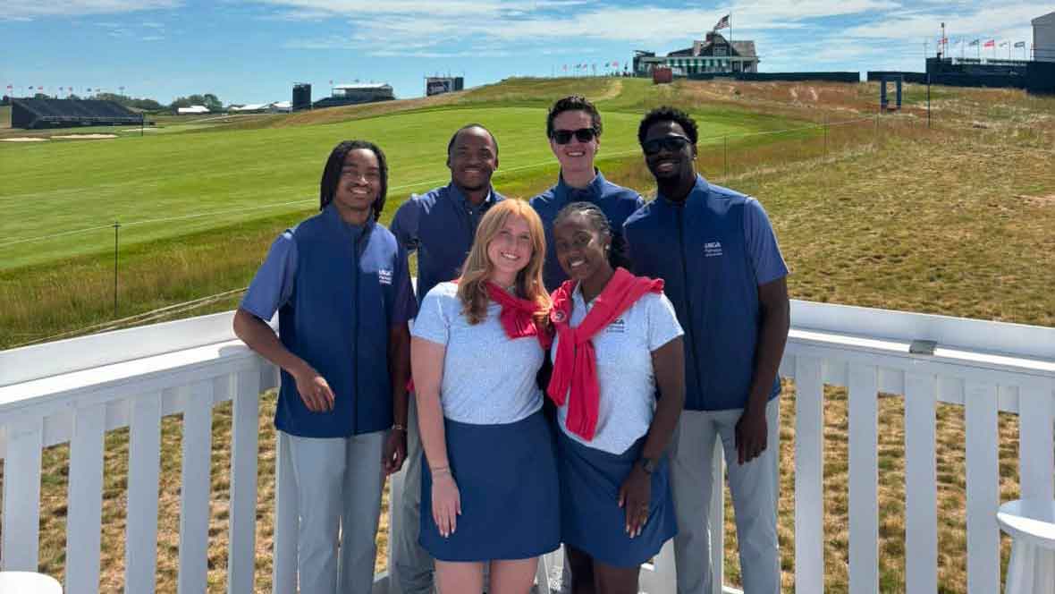 Six people wearing matching blue and white outfits stand and smile together on a white deck overlooking a grassy golf course on a sunny day.