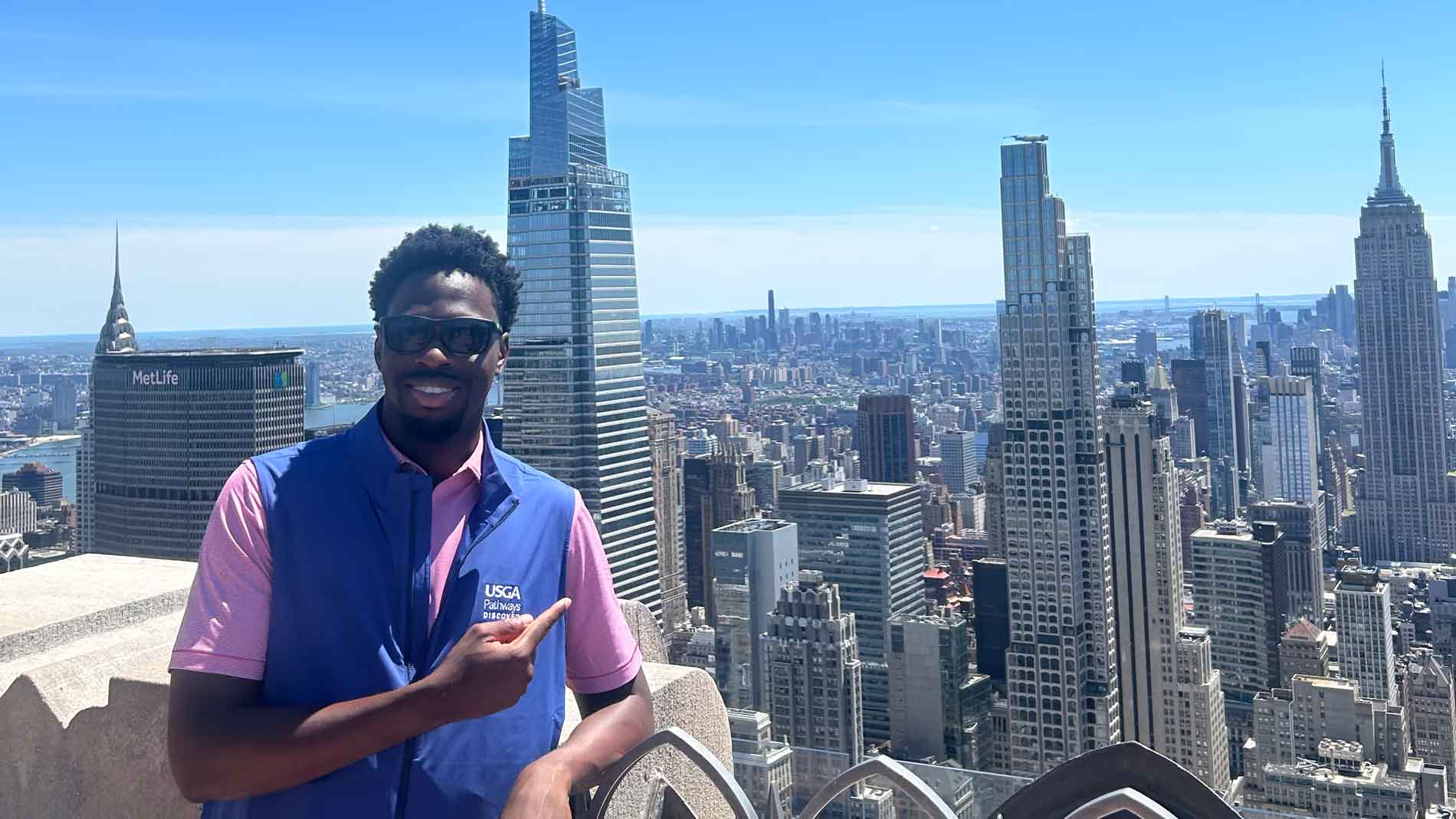 A man in sunglasses and a blue vest smiles and points with one hand on a rooftop observation deck, with the New York City skyline, including the Empire State Building, in the background under a clear blue sky.